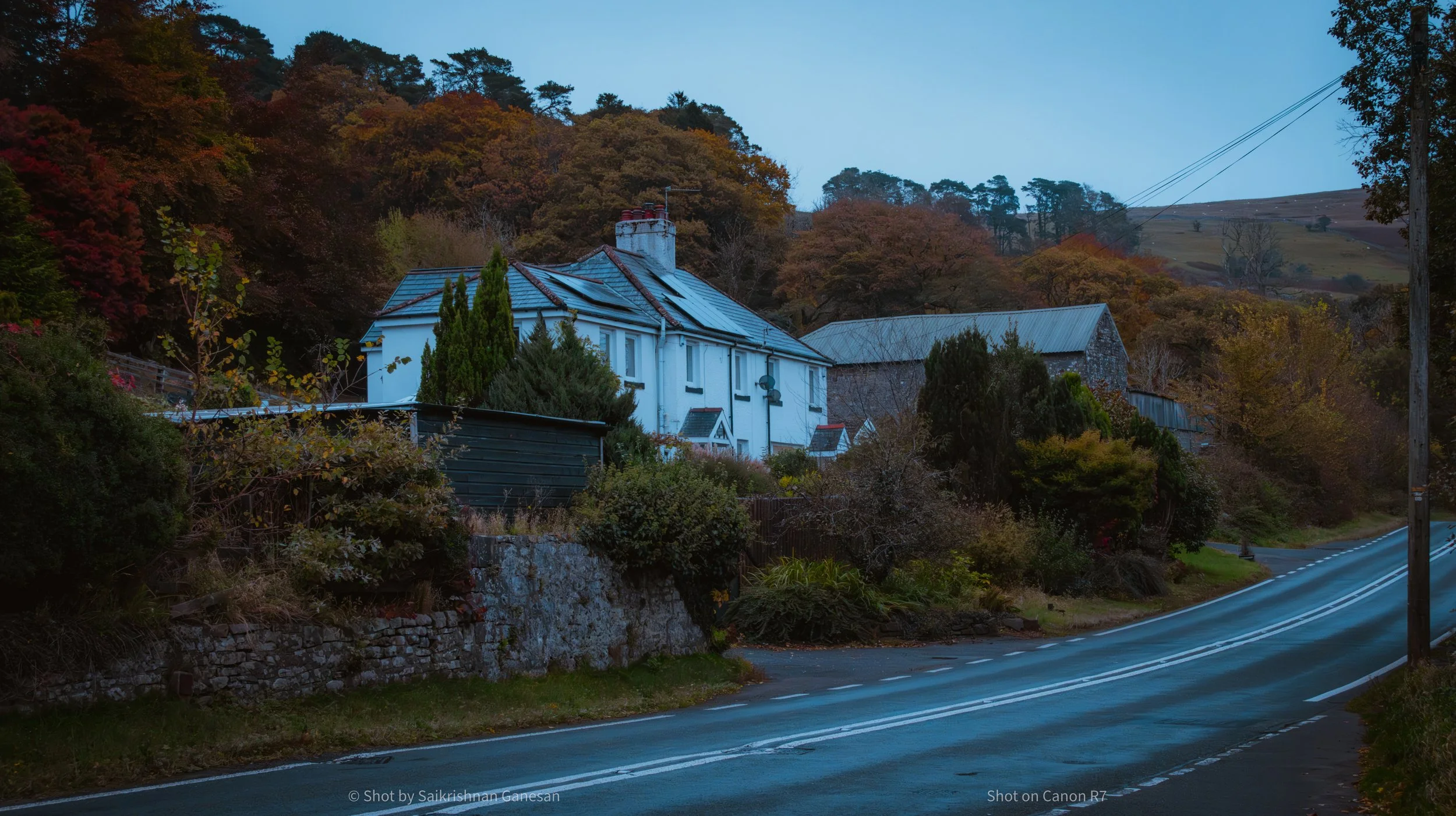 A winding road beside a house on a hillside with trees displaying fall foliage in the background.