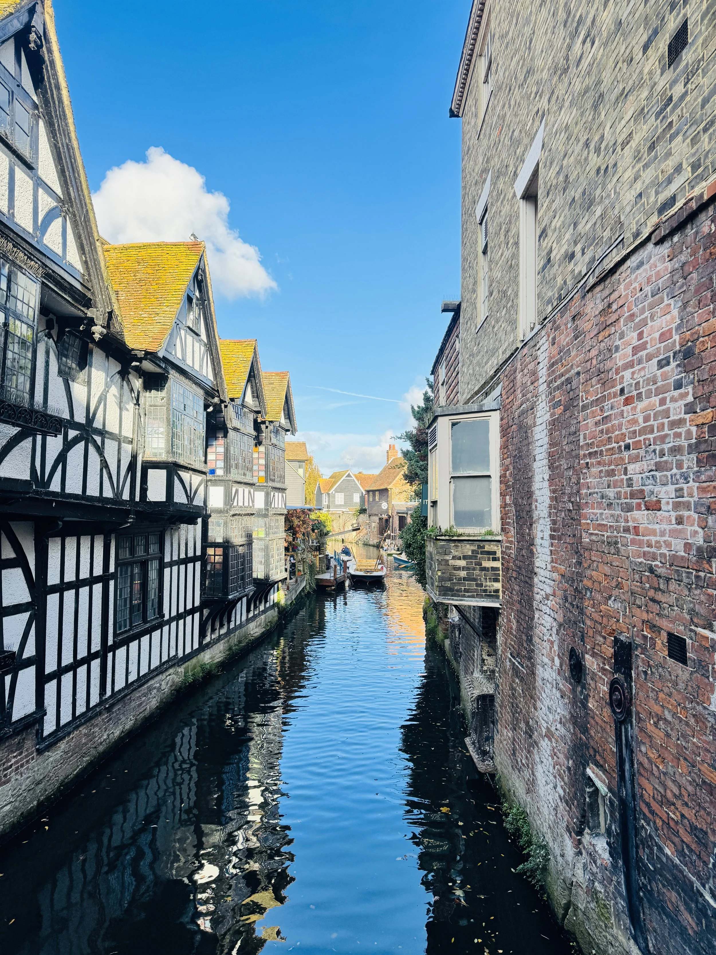 A narrow canal surrounded by old buildings with brick and timber facades, some with yellow moss-covered rooftops, with boats docked along the sides under a blue sky with clouds.
