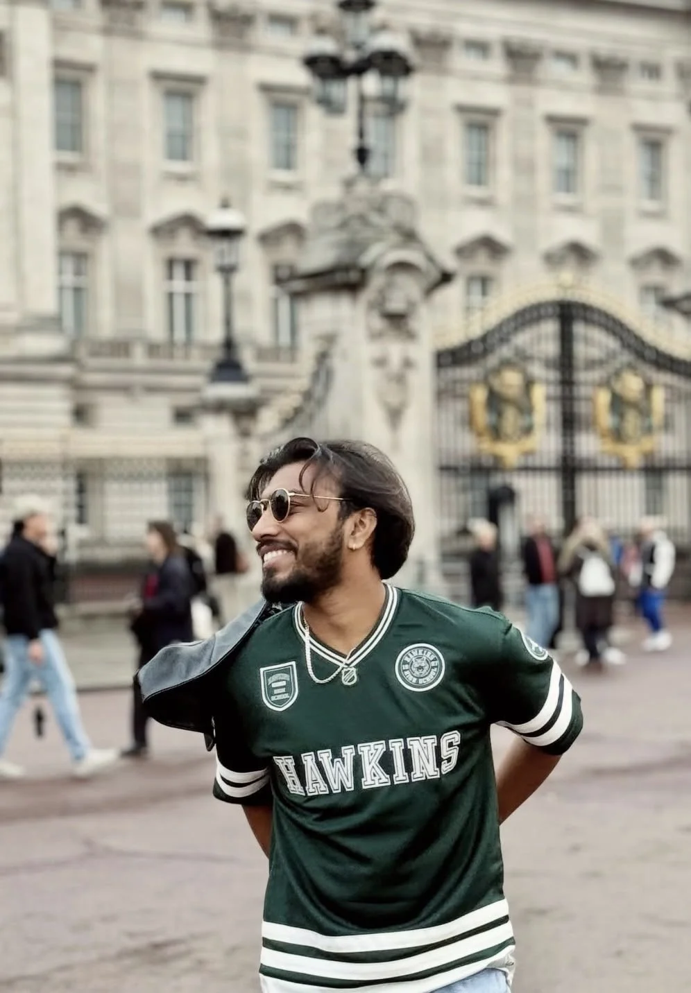 A smiling man wearing sunglasses and a green sports jersey with 'Hawkins' written on it, standing outdoors in front of an ornate historic building with a gated entrance, with people walking in the background.