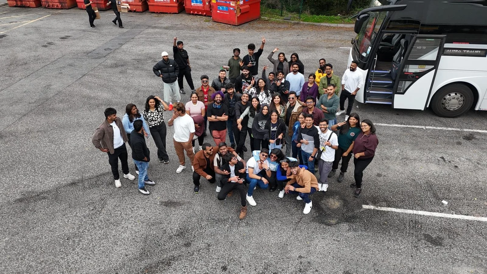 A large group of people standing in a parking lot, posing for a photo, with a white bus to the right and red dumpsters in the background.