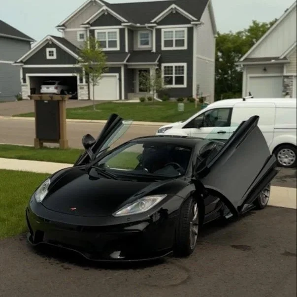 A black sports car with butterfly doors open parked on a driveway in front of a suburban house with gray and white siding. A white van and a gray house are visible in the background.