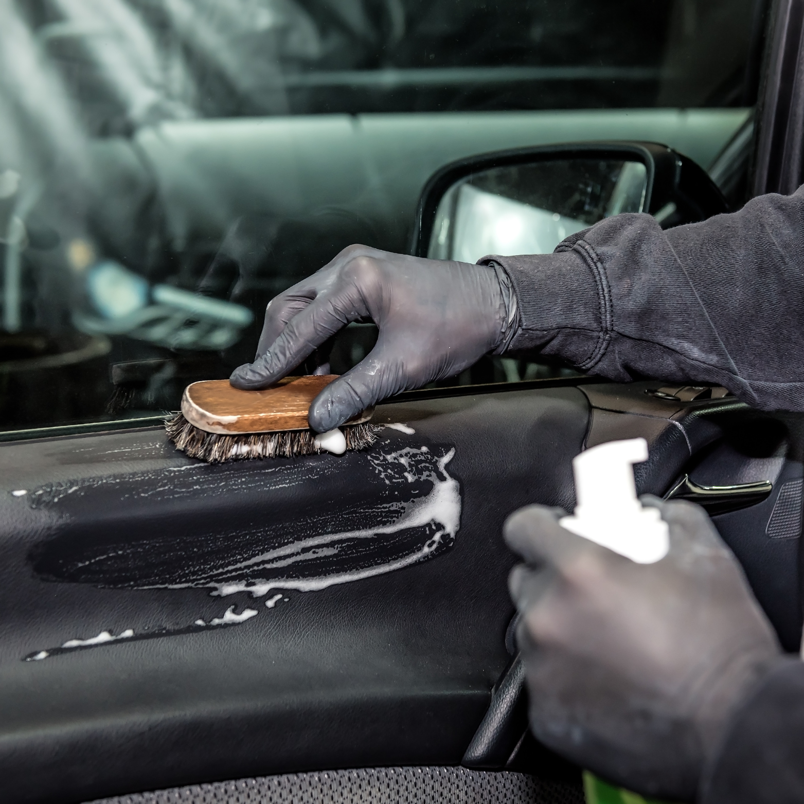Person cleaning a vehicle's dashboard with a brush and spray bottle, wearing black gloves.
