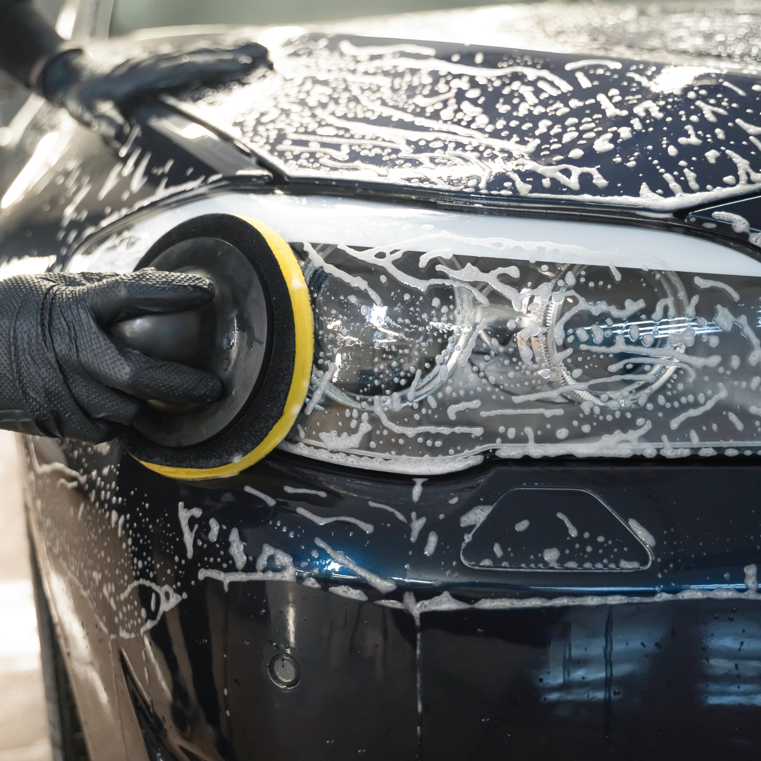 A person wearing black gloves is polishing the headlight of a black car with a yellow and black buffer pad, soap and water are visible on the car.