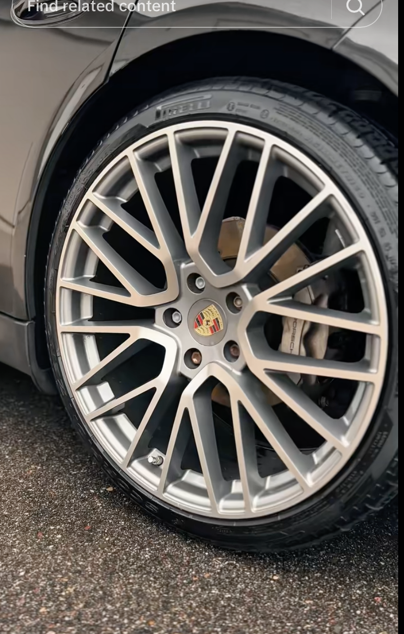 Close-up of a Porsche wheel with a silver alloy rim, black brake caliper, and Pirelli tire on a textured asphalt surface.