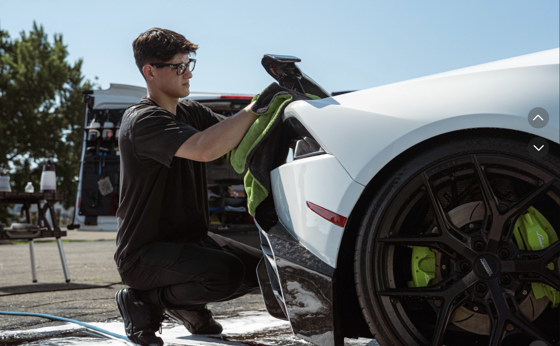 A young man wearing glasses and black clothing is cleaning or polishing a white sports car with a green towel. The car has black rims and green brake calipers. There is a work van in the background with its back doors open, revealing tools and supplies. The scene appears to be outdoors on a paved area under a clear blue sky.