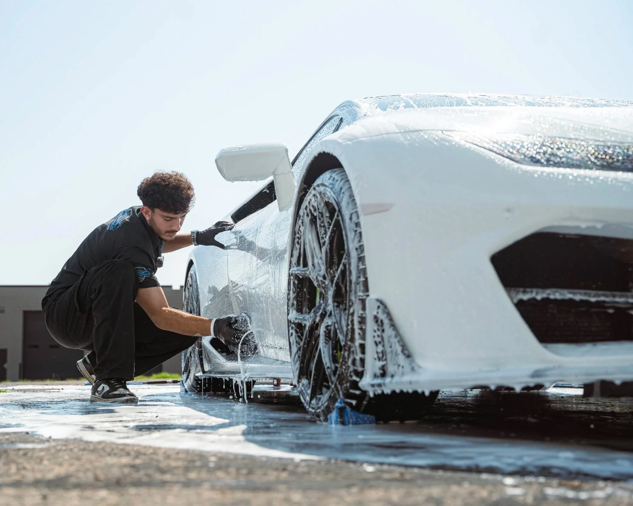 A man washing a white sports car with foam outdoors on a sunny day.