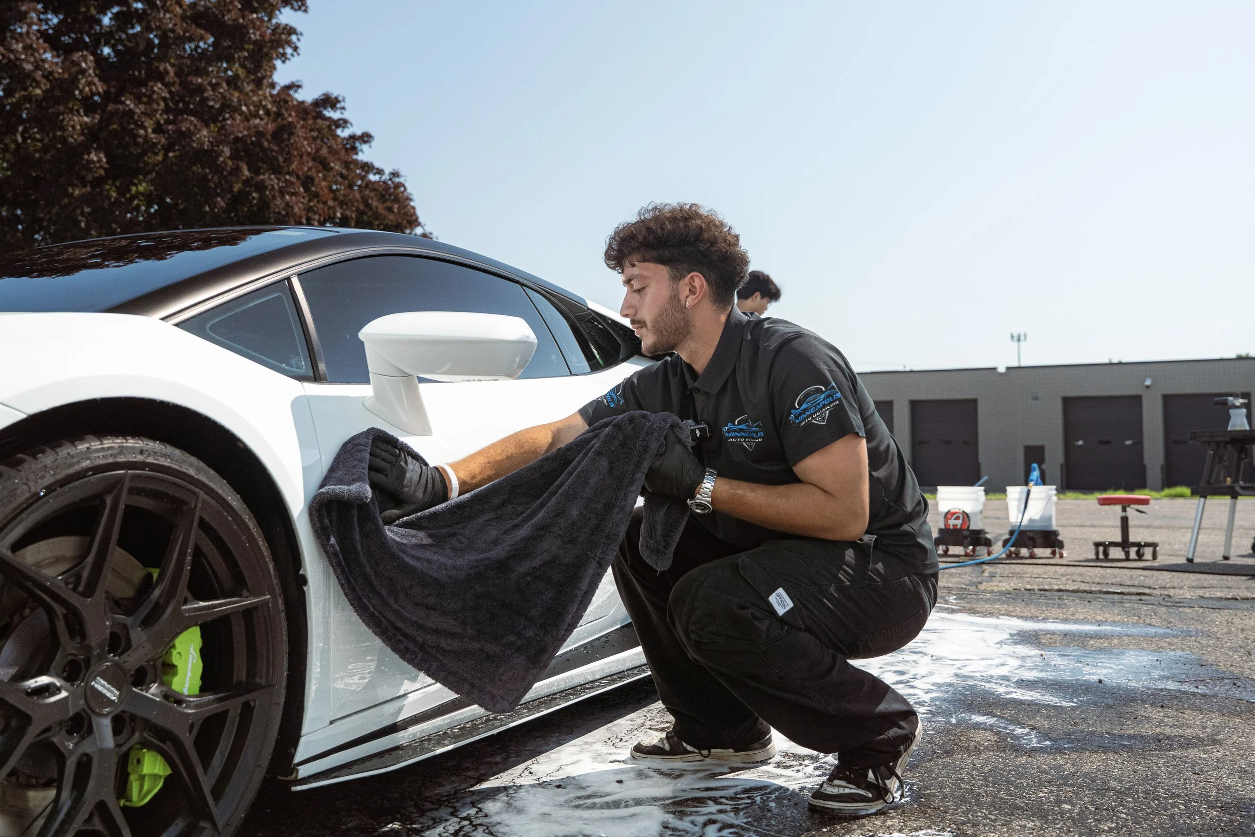A man in black uniform cleaning the side of a white sports car outside on a sunny day with towels, cleaning supplies, and tools visible in the background.