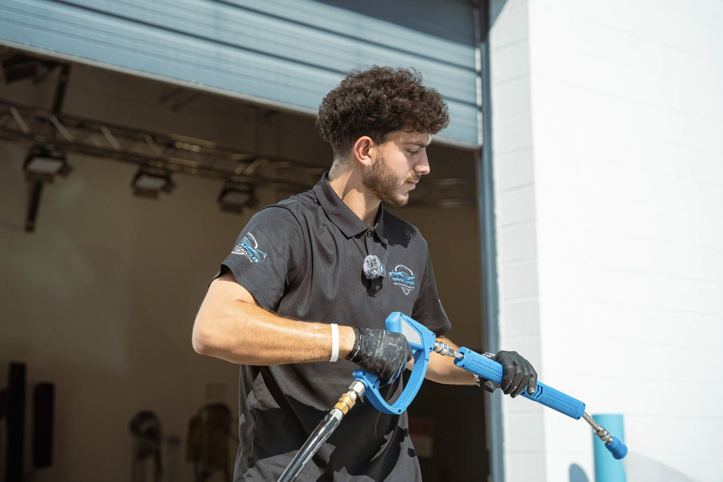 A man in a black polo shirt with a logo, wearing black gloves, holding and operating a blue pressure washer outdoors.