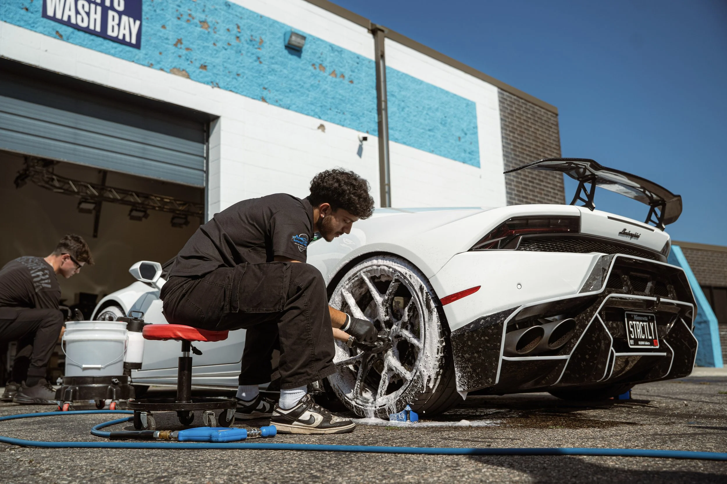 A man washing a white Lamborghini sports car with soap and water outside a garage, with another person in the background.