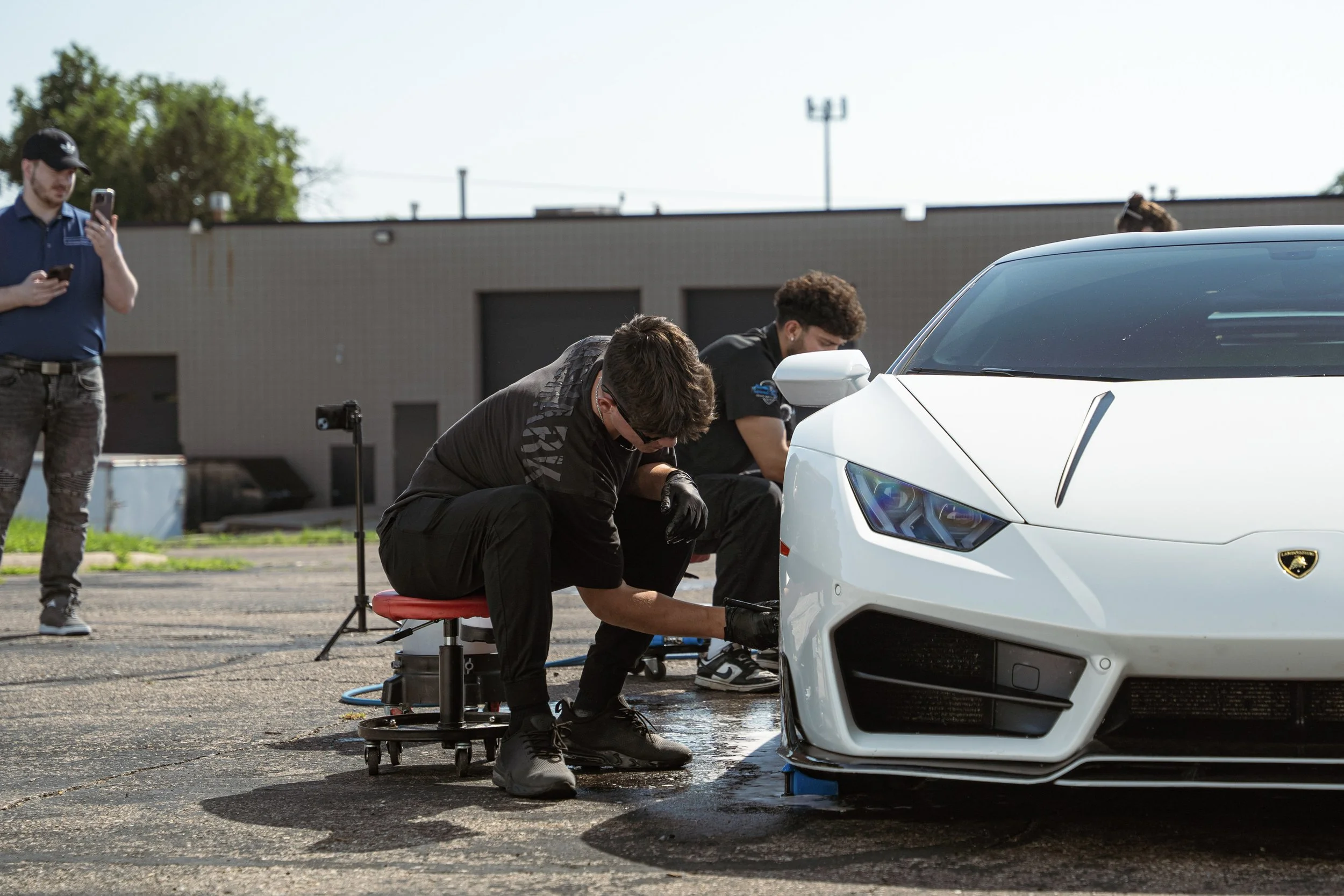 Two mechanics inspecting a white sports car outdoors with a man recording on his phone and another person sitting nearby.