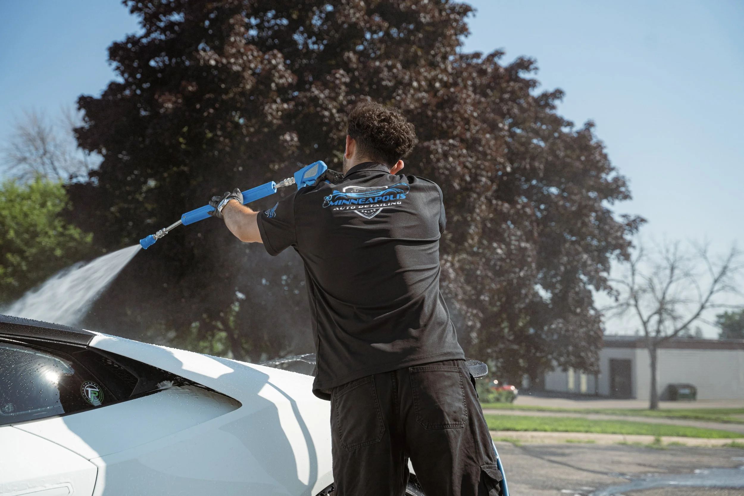 A person washing a white sports car with a high-pressure water spray. The person is wearing a black t-shirt with blue lettering and black shorts, standing outdoors on a sunny day.