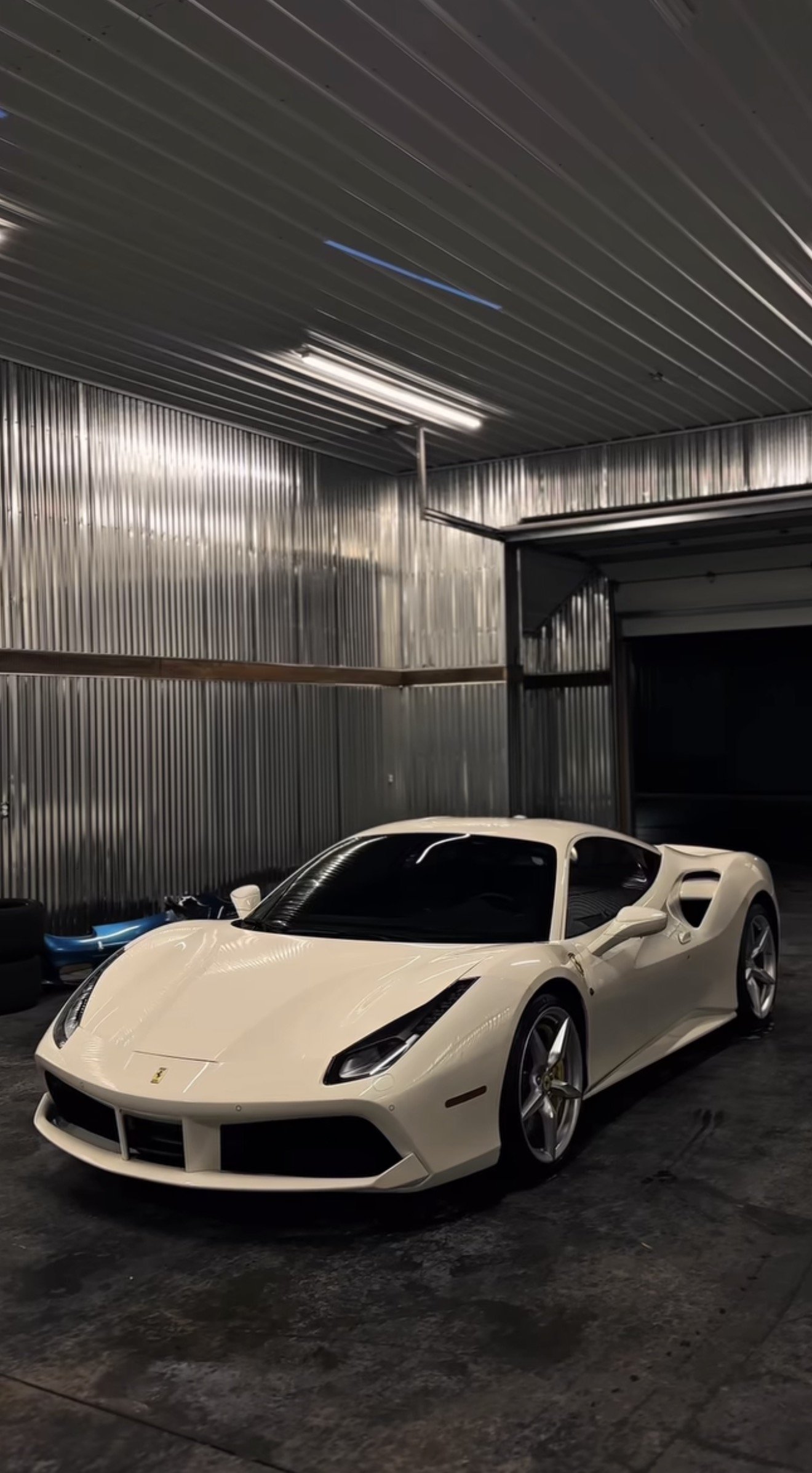 A white luxury sports car, resembling a Ferrari LaFerrari, parked inside a metallic industrial garage with corrugated steel walls and ceiling.