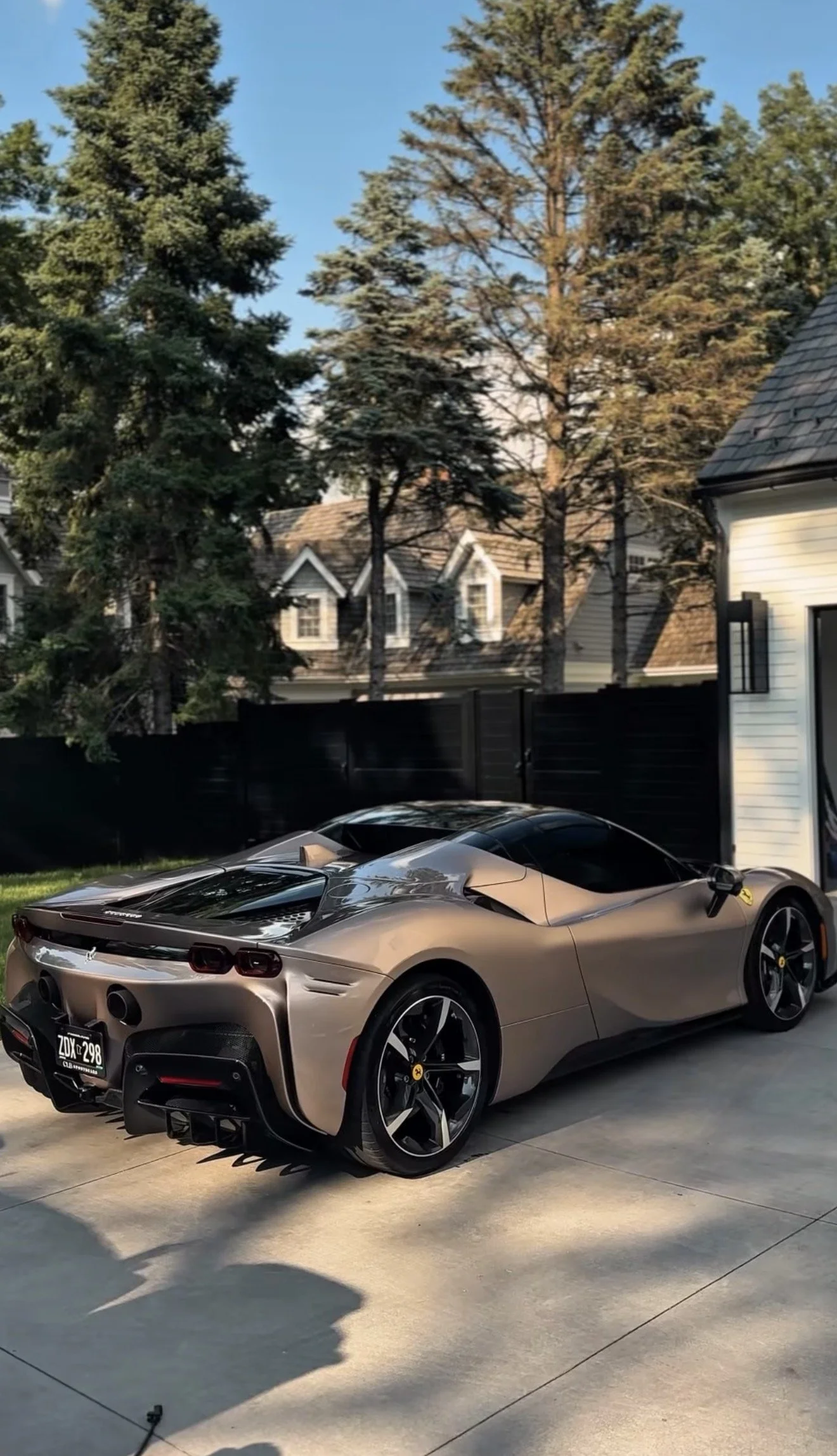 A metallic grey Ferrari sports car parked on a concrete driveway in front of a white house with black fences, surrounded by tall trees against a clear blue sky.