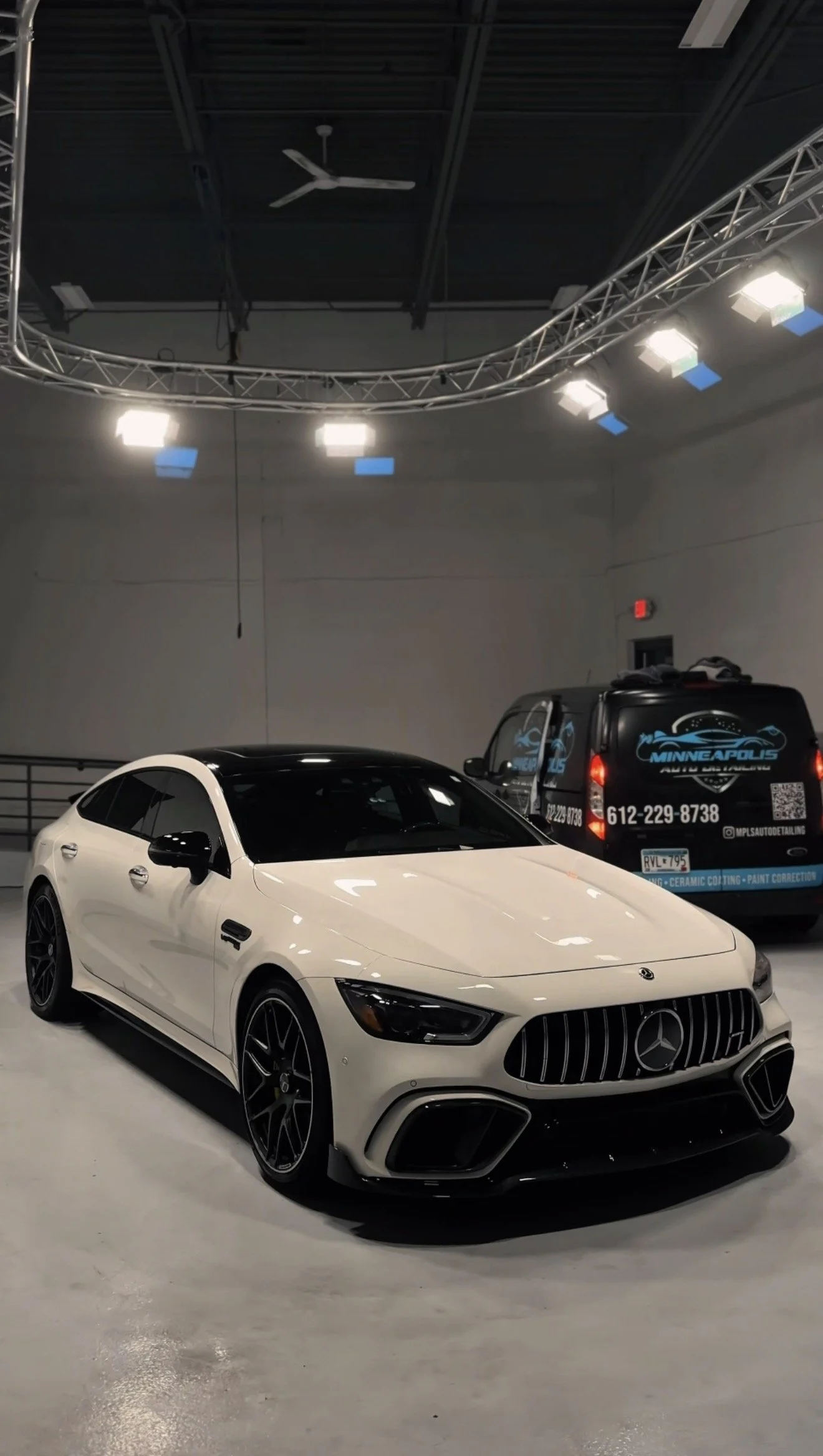 A white Mercedes-Benz car with black accents parked inside a garage with bright overhead lights and another vehicle in the background with dealership signage.