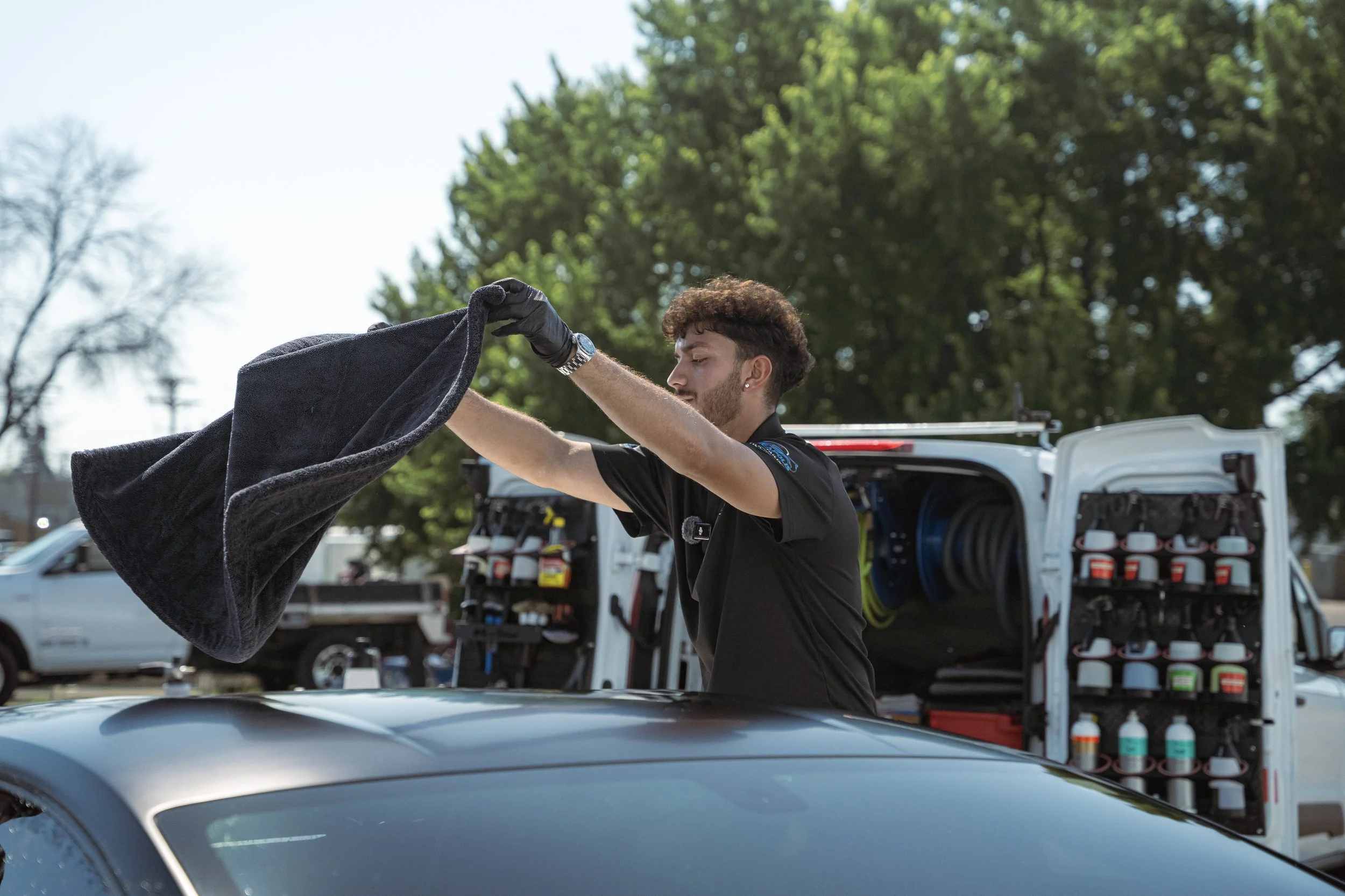 A man wearing black gloves and a black shirt is placing a black towel on the roof of a silver car. In the background, a service van with various bottles and equipment is parked under a tree with green leaves.