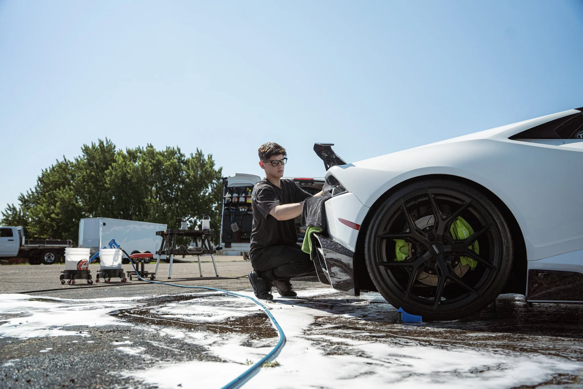 Man washing a white sports car outdoors on a sunny day, with equipment and trees in the background.