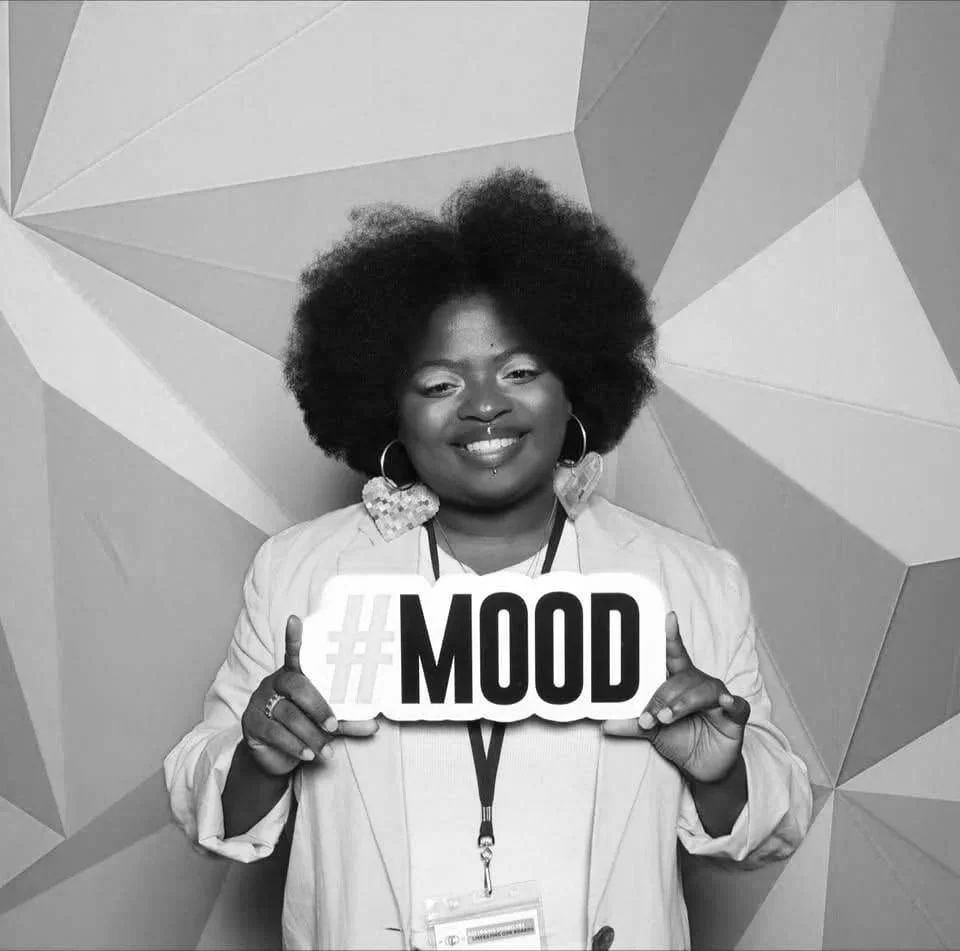 Erica smiling and holding a sign that reads '#MOOD' in front of a geometric-patterned background.