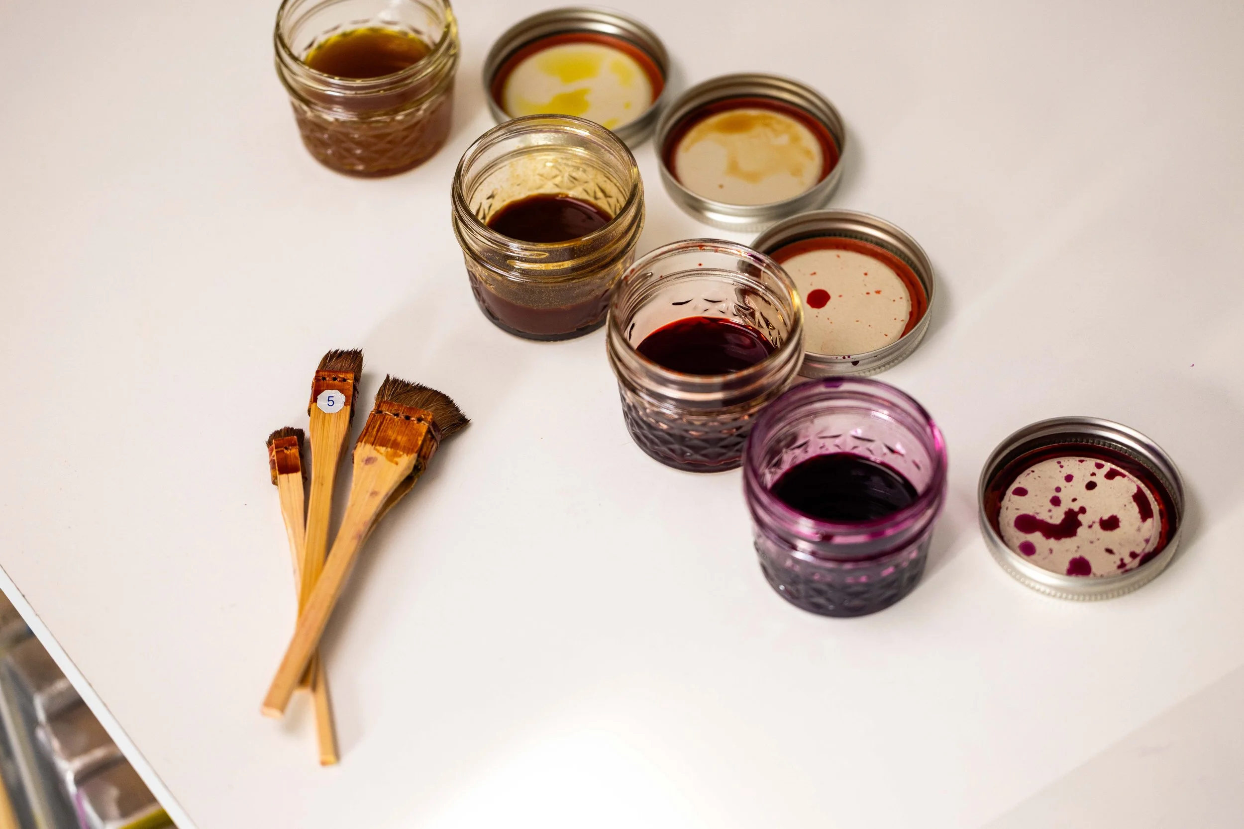 Six small jars containing various colors of ink with their lids off, arranged in a semi-circle on a white surface. Four paintbrushes with wooden handles and brown bristles rest on the surface to the left of the jars.