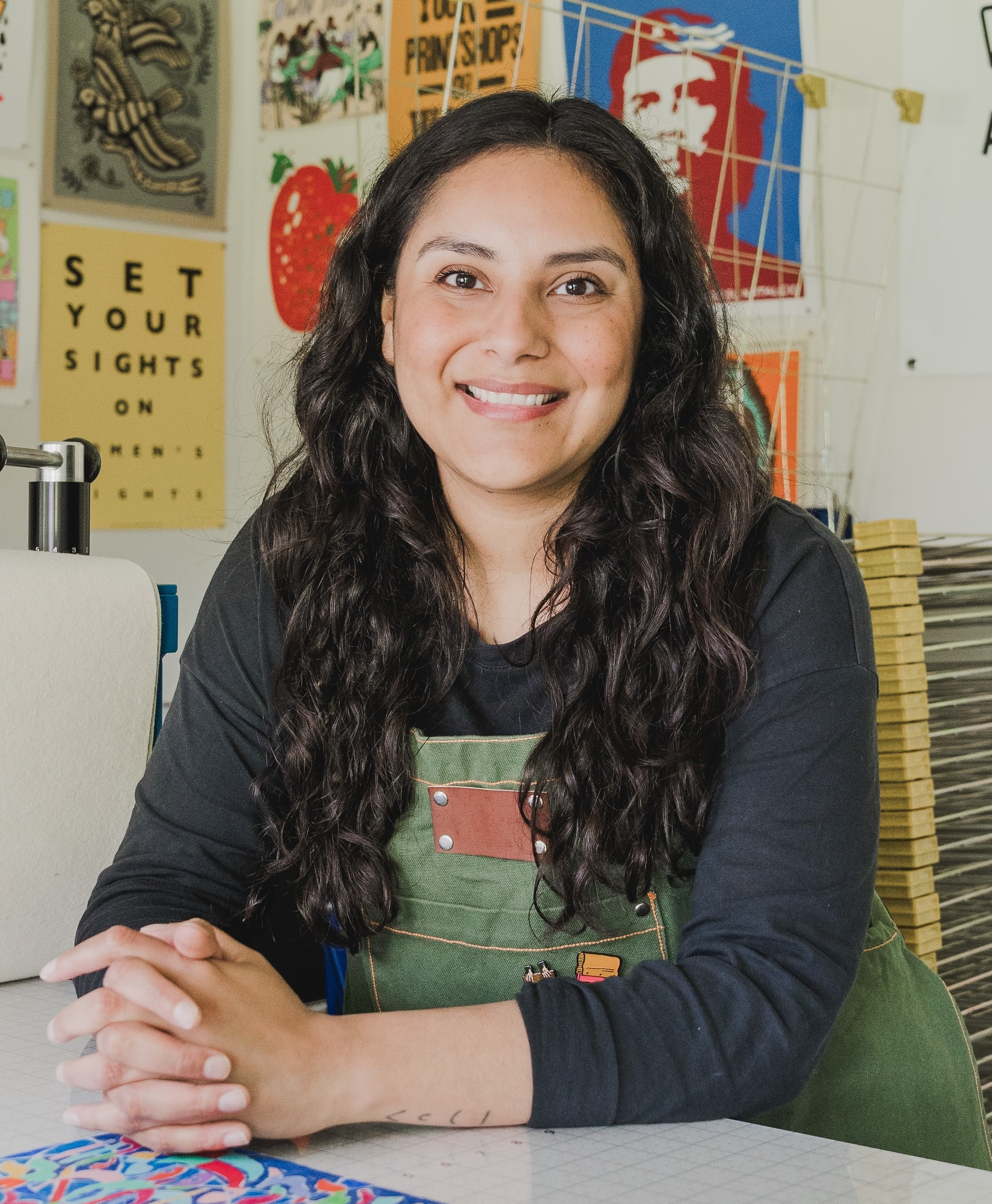 Woman with long curly dark hair smiling, sitting at a table in an arts and crafts room with colorful posters and artwork in the background.