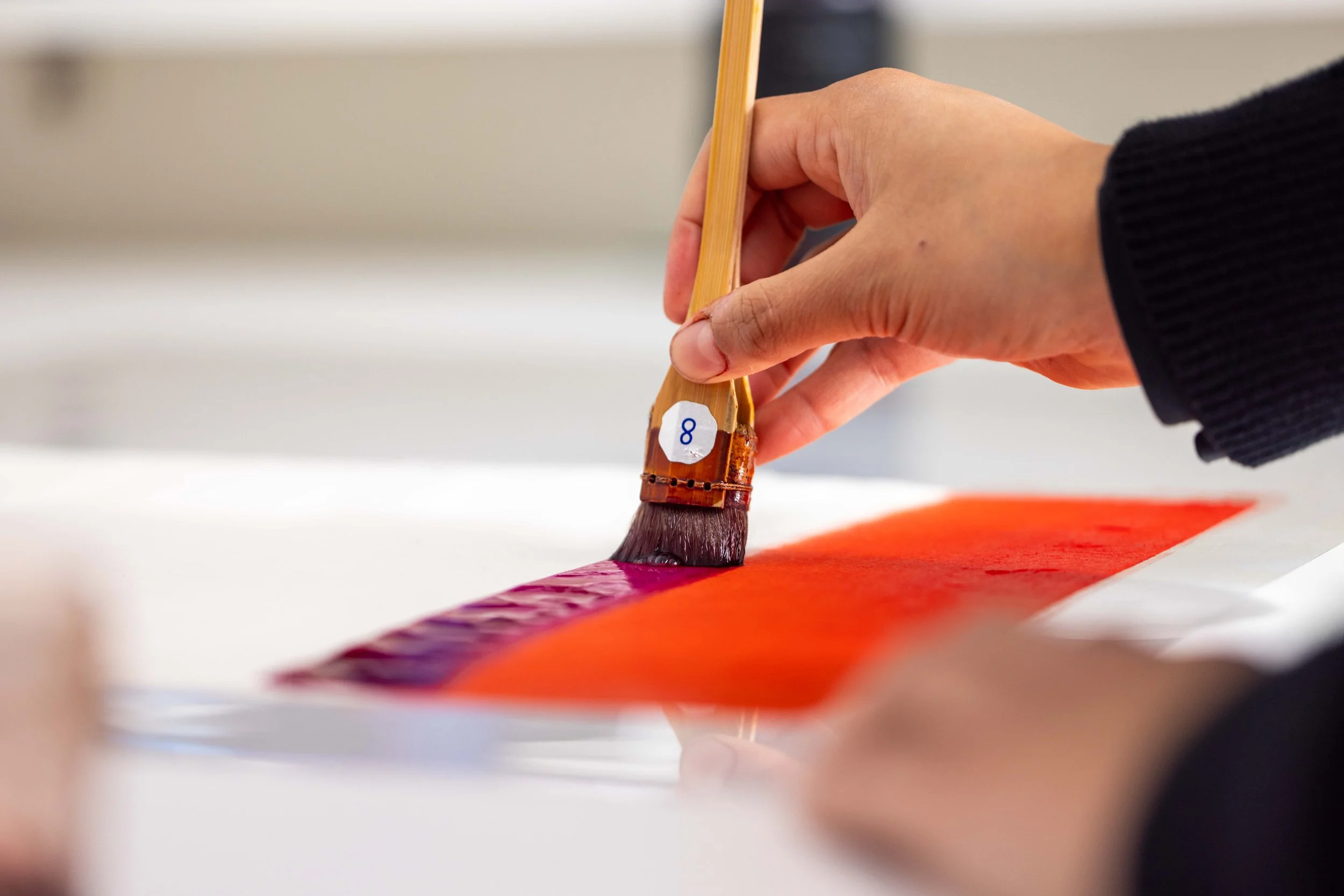 Hand holding a paintbrush, painting with red, purple, and orange colors on paper. 