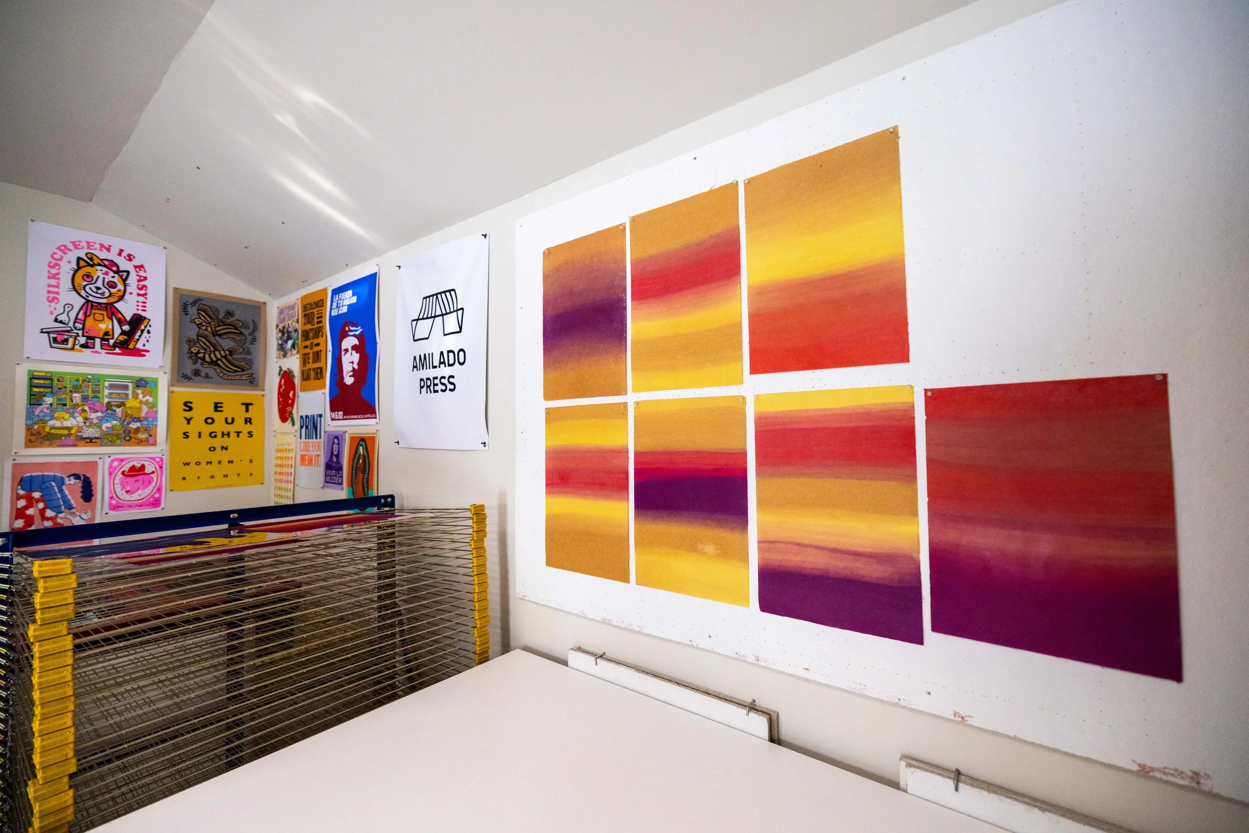 Gallery wall with colorful posters and abstract art, including illustrations, text designs, and a sunset color gradient artwork, in an art studio or exhibition space.