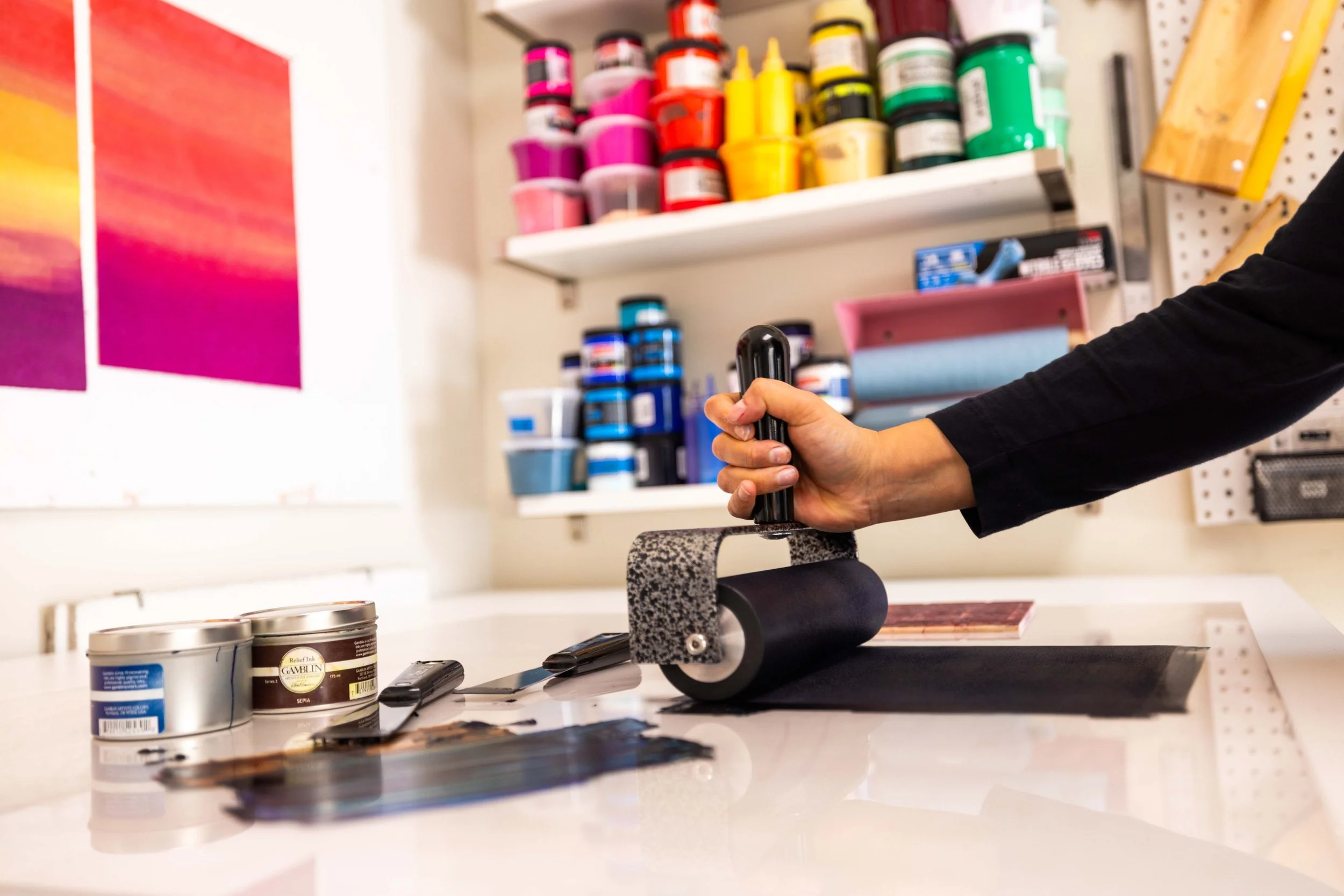 A person using a paint roller to apply black paint to a surface in an art or craft workspace. The workspace has shelves with colorful paints and supplies in the background.