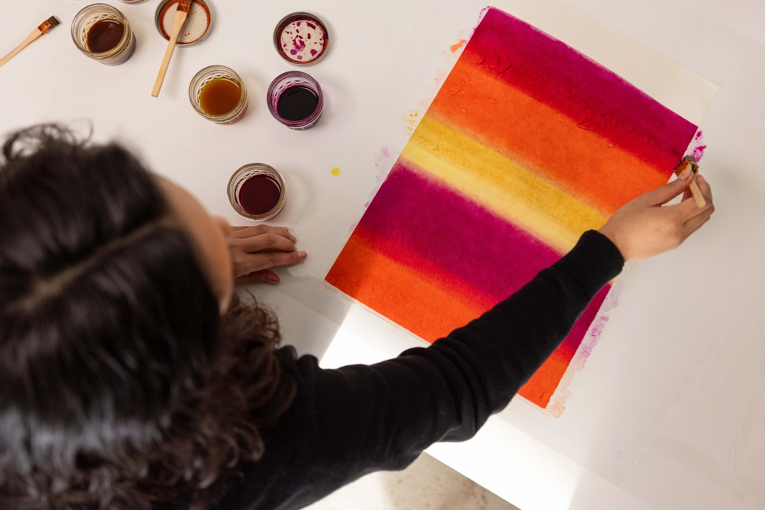 Person using a sponge to create an abstract watercolor painting with vibrant pink, orange, yellow, and purple stripes, surrounded by small jars of watercolor paints on a white table.