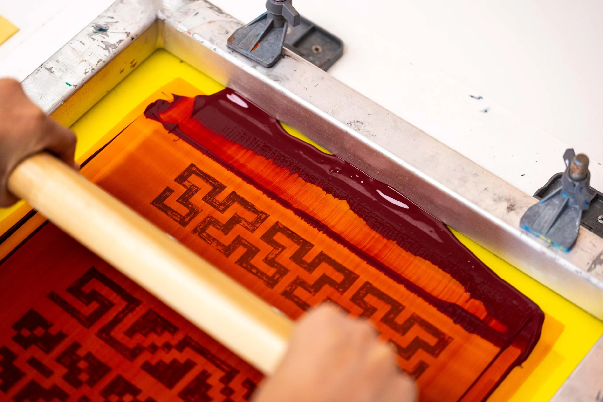 A person using a screen printing frame to print a red and orange design with geometric patterns.