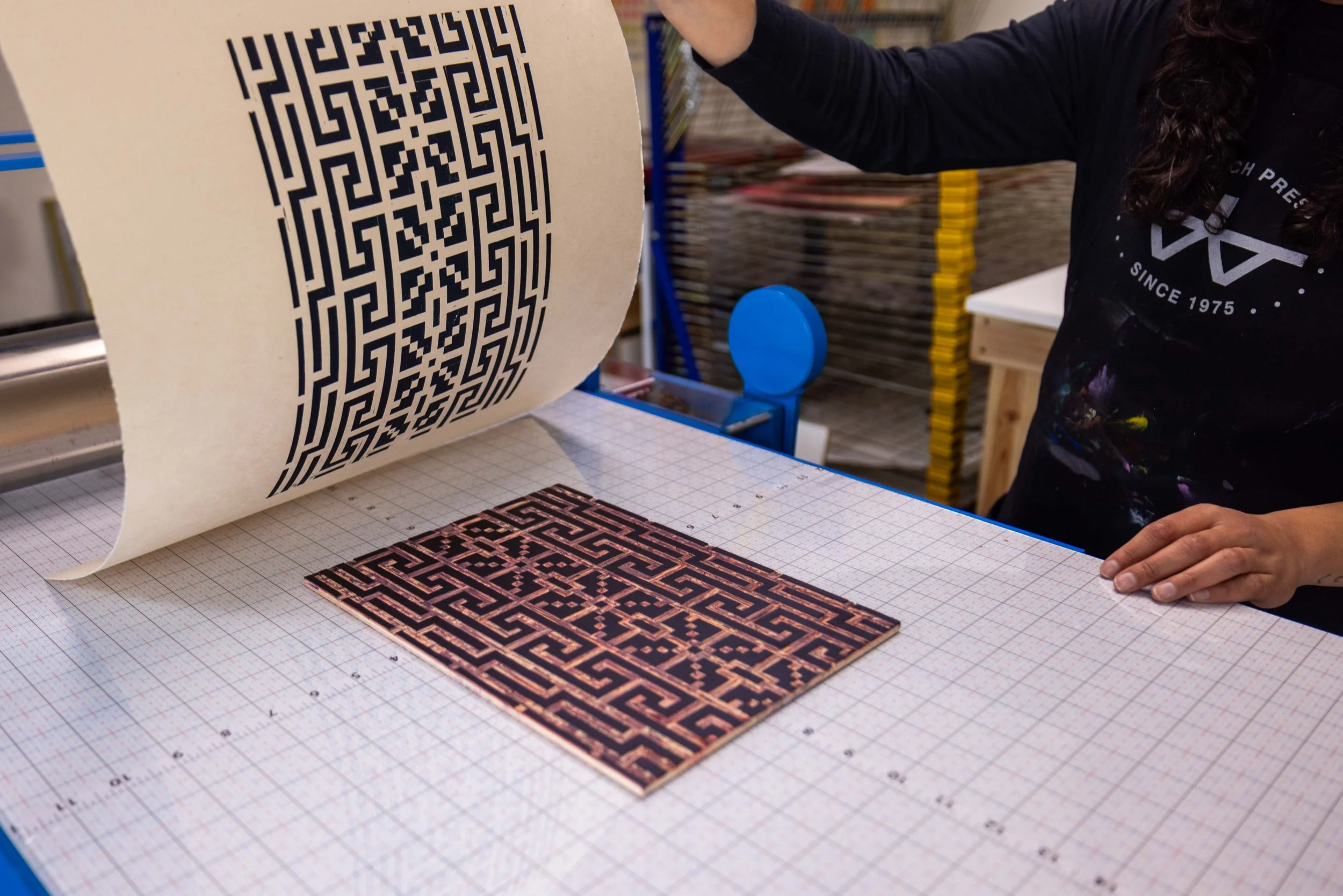 A person holding a large printed print with a maze-like pattern, on a grid-lined table in a workspace.