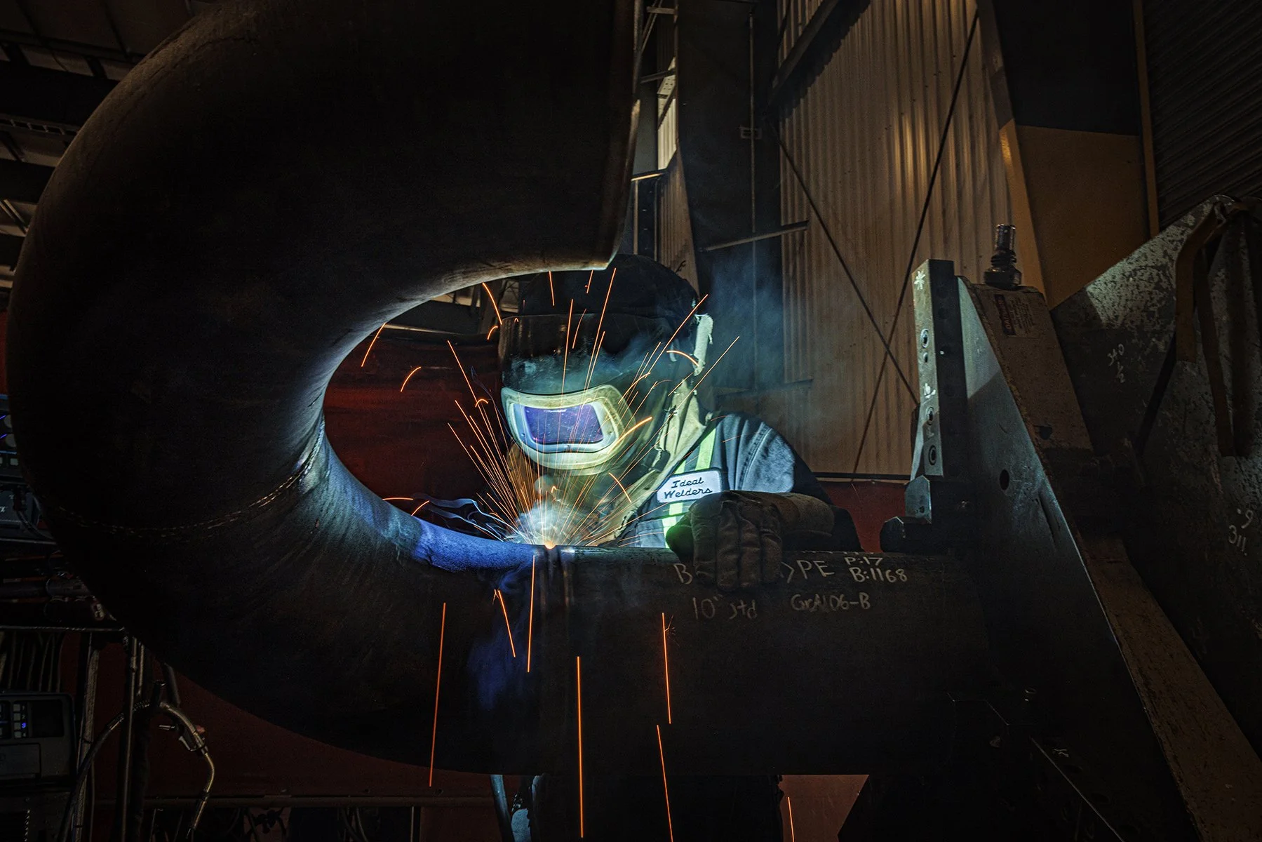 A welder wearing protective gear welding a large metal pipe, with sparks flying in a workshop.