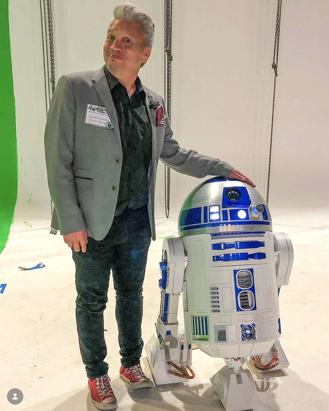 A man in a gray blazer and black shirt standing next to a life-sized model of R2-D2 from Star Wars.