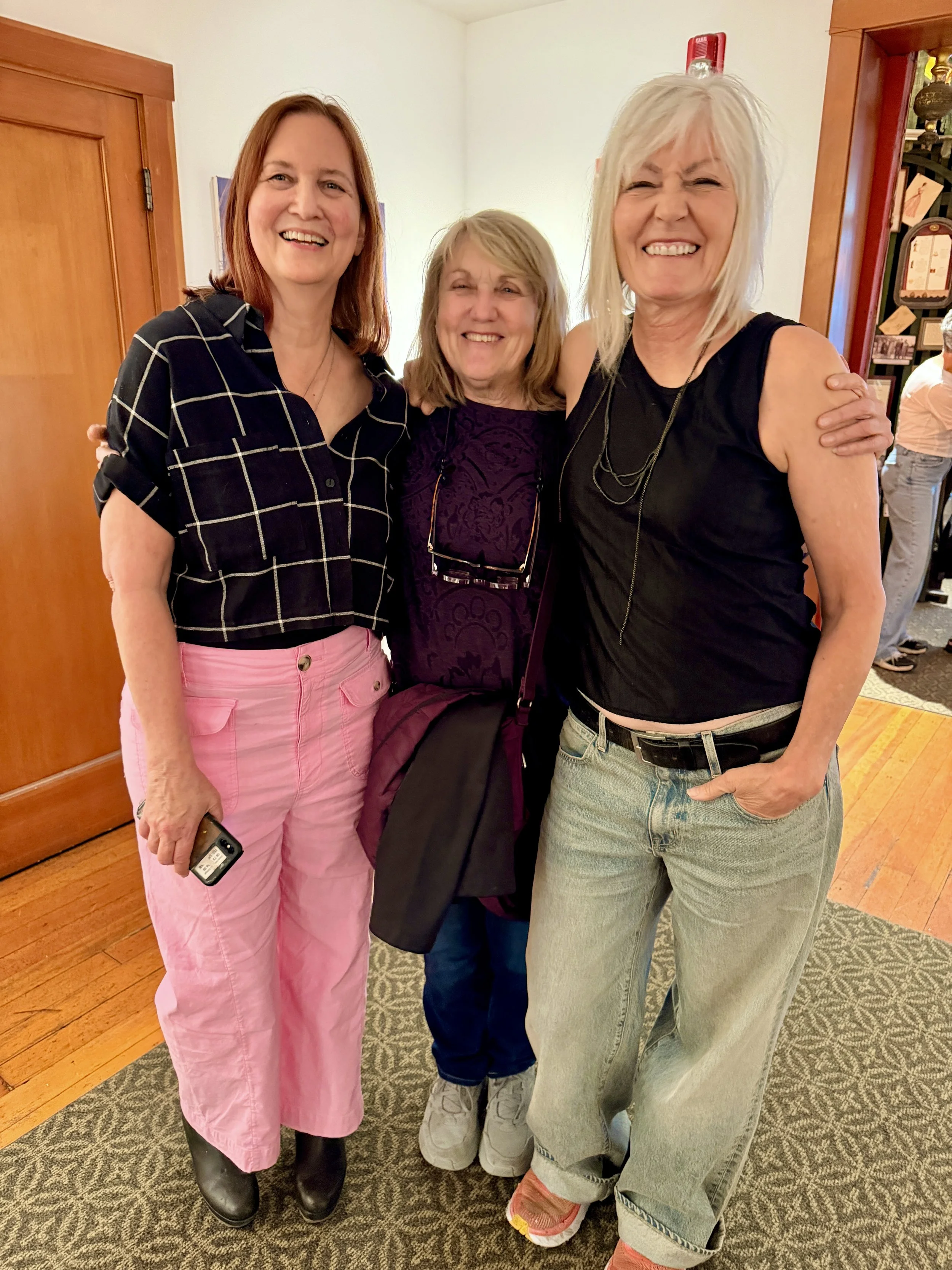 Three women standing together indoors, smiling at the camera. The woman on the left has red hair, is wearing a black and white checked shirt and pink pants. The woman in the middle has blonde hair, is wearing a purple top and glasses hanging from her neck. The woman on the right has light blonde hair, is wearing a black sleeveless top and light jeans, with her hand on the shoulder of the woman in the middle.