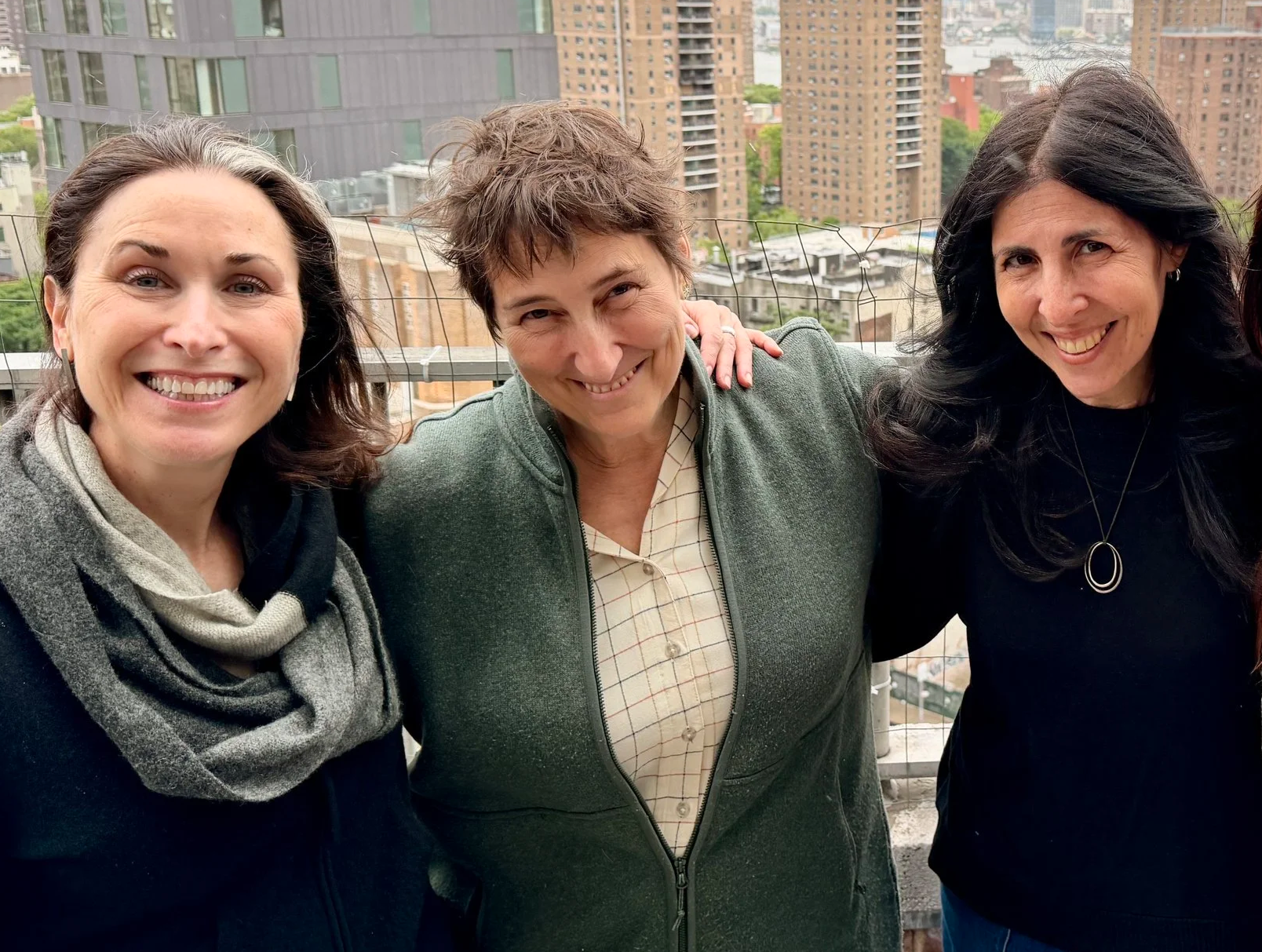 Three women standing close together on a rooftop with city buildings in the background, smiling at the camera.
