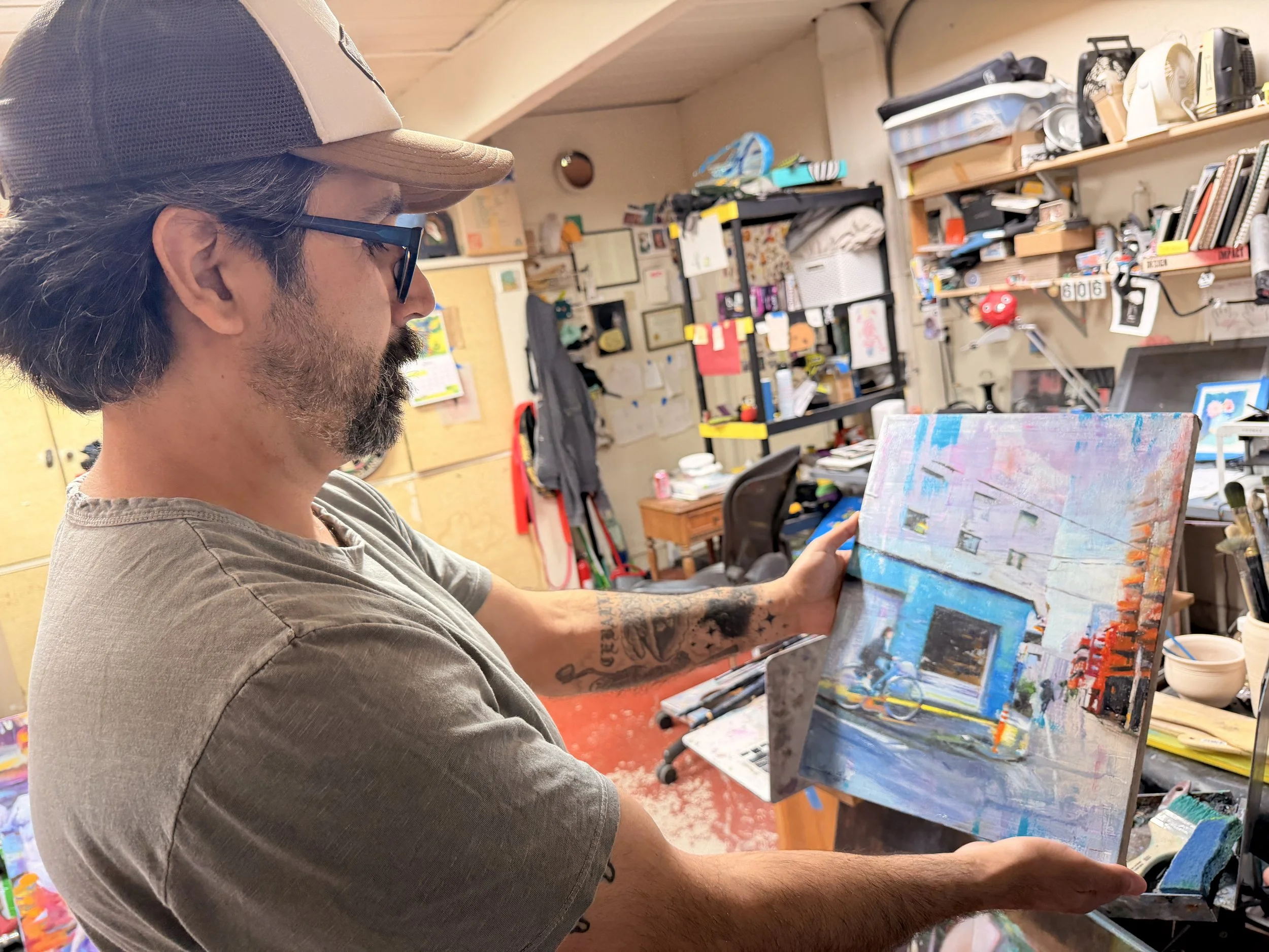 A man wearing a baseball cap and glasses is holding a painting on an easel in an art studio. The studio has shelves filled with art supplies, books, and various items, with a cluttered workspace in the background.