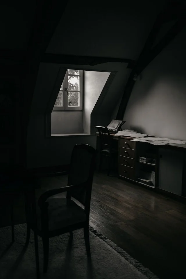 A dimly lit attic room with a window, a wooden desk with papers, and a chair.