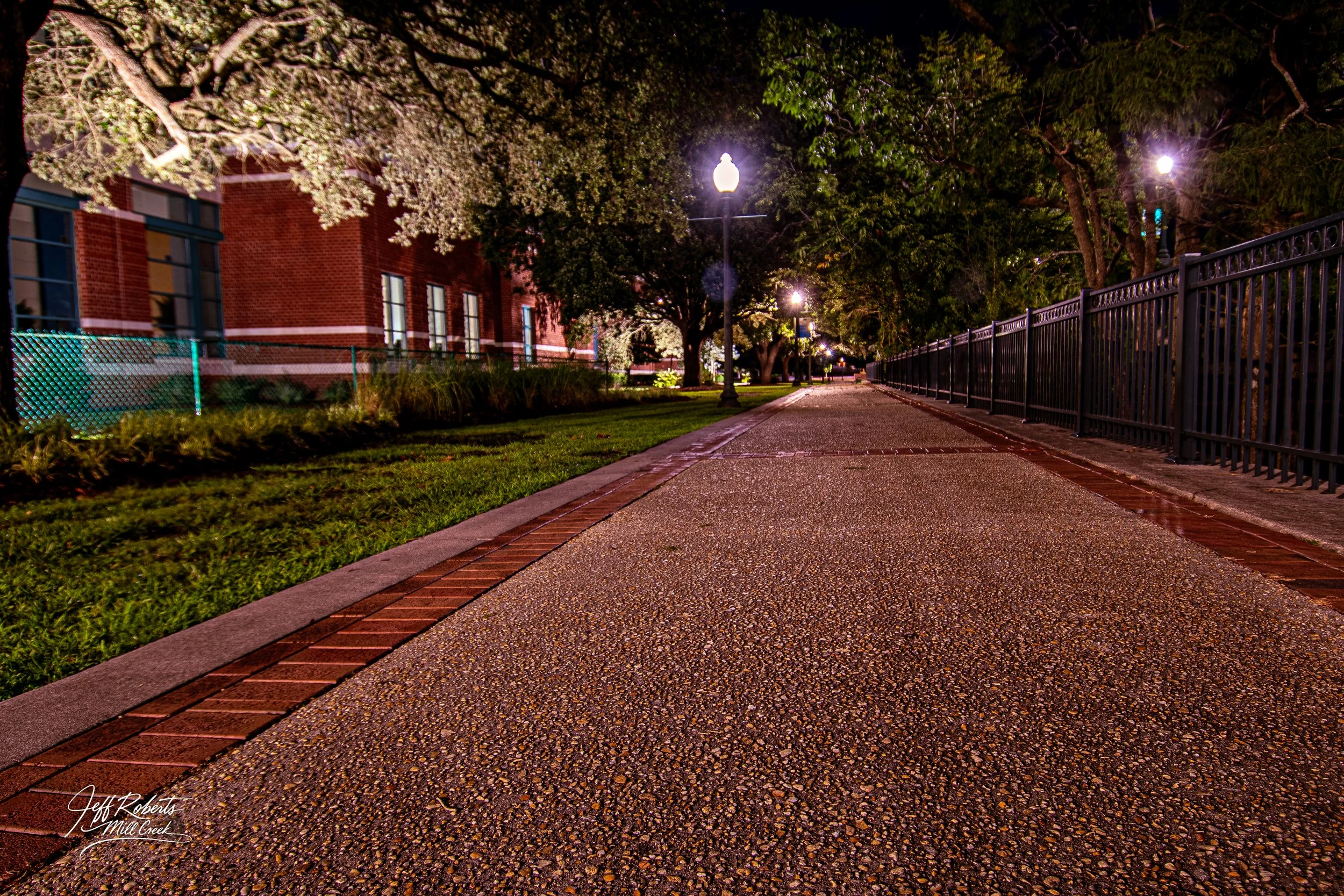 Nighttime view of a sidewalk with streetlights, trees, and a fence along one side, and a building with windows on the other side.