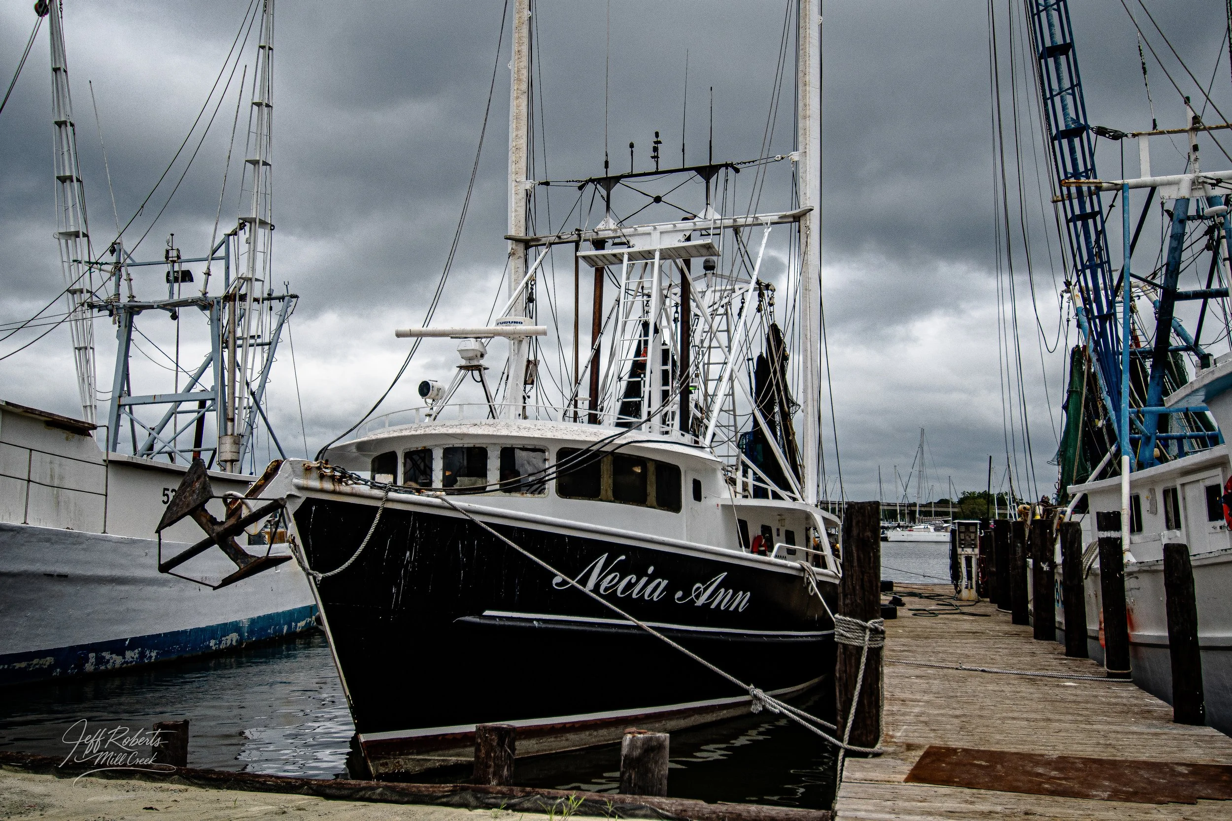 A black and white boat named 'Necia Ann' docked at a marina under dark, cloudy sky, surrounded by several other boats and anchored to wooden piers.