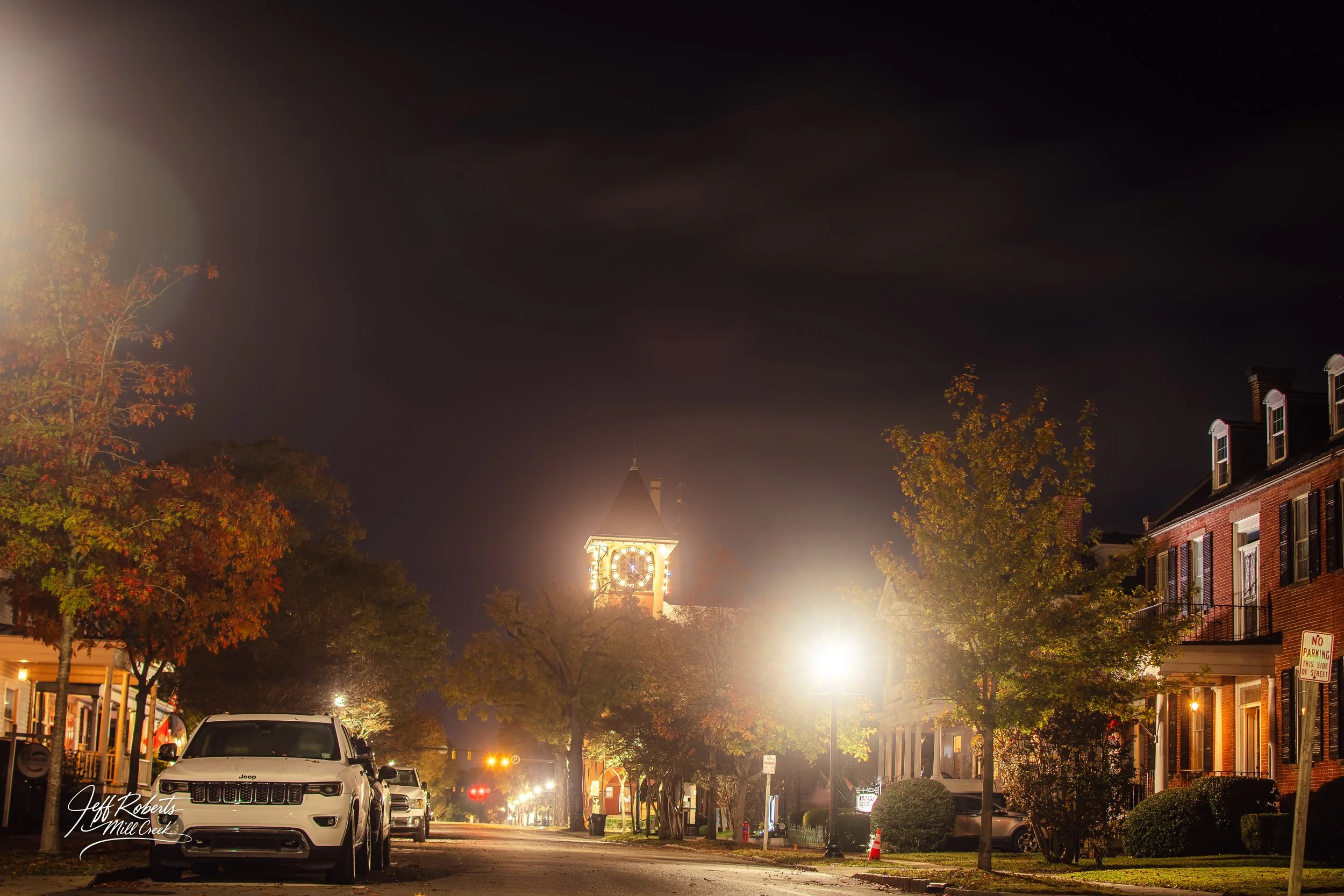 Nighttime street view with historic clock tower illuminated in the background, trees with autumn foliage, parked cars, and brick buildings lining the street.