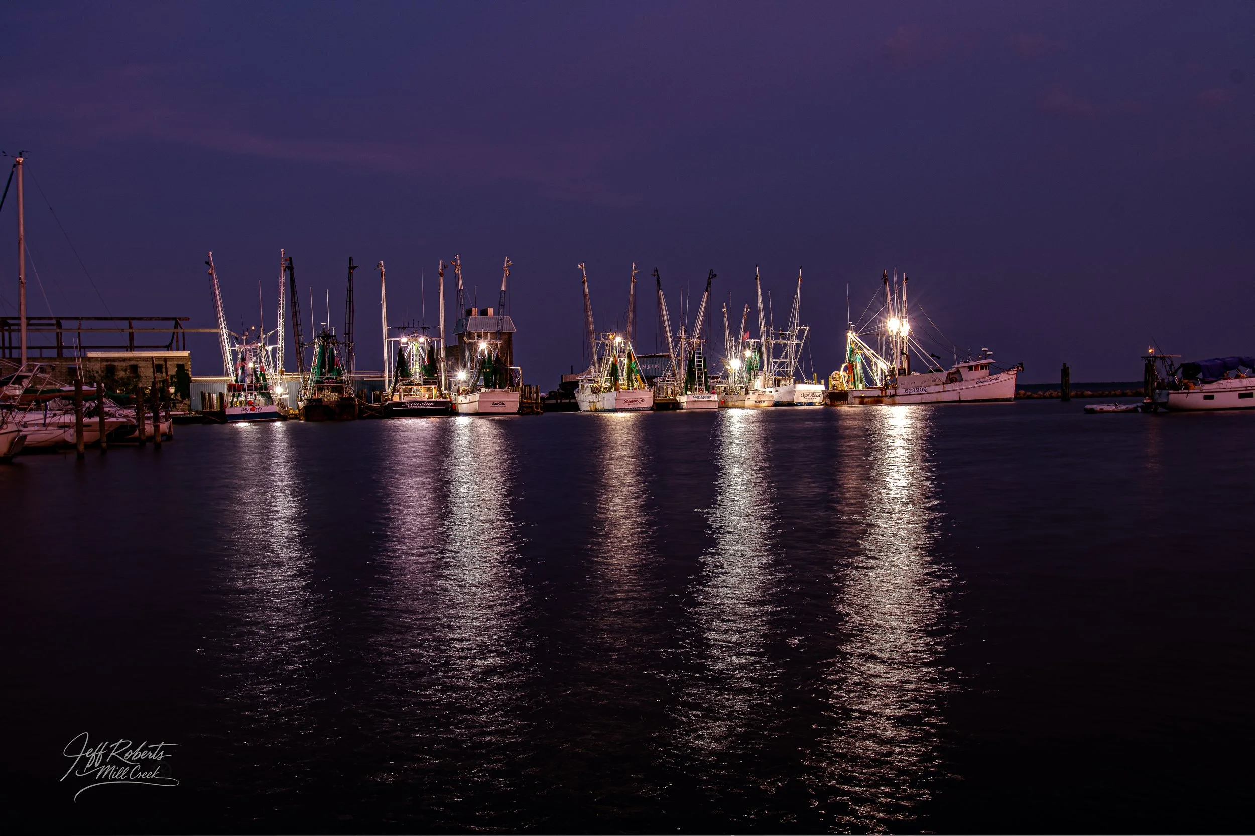 Nighttime view of docked boats at a marina with reflections on calm water, illuminated by bright boat lights.