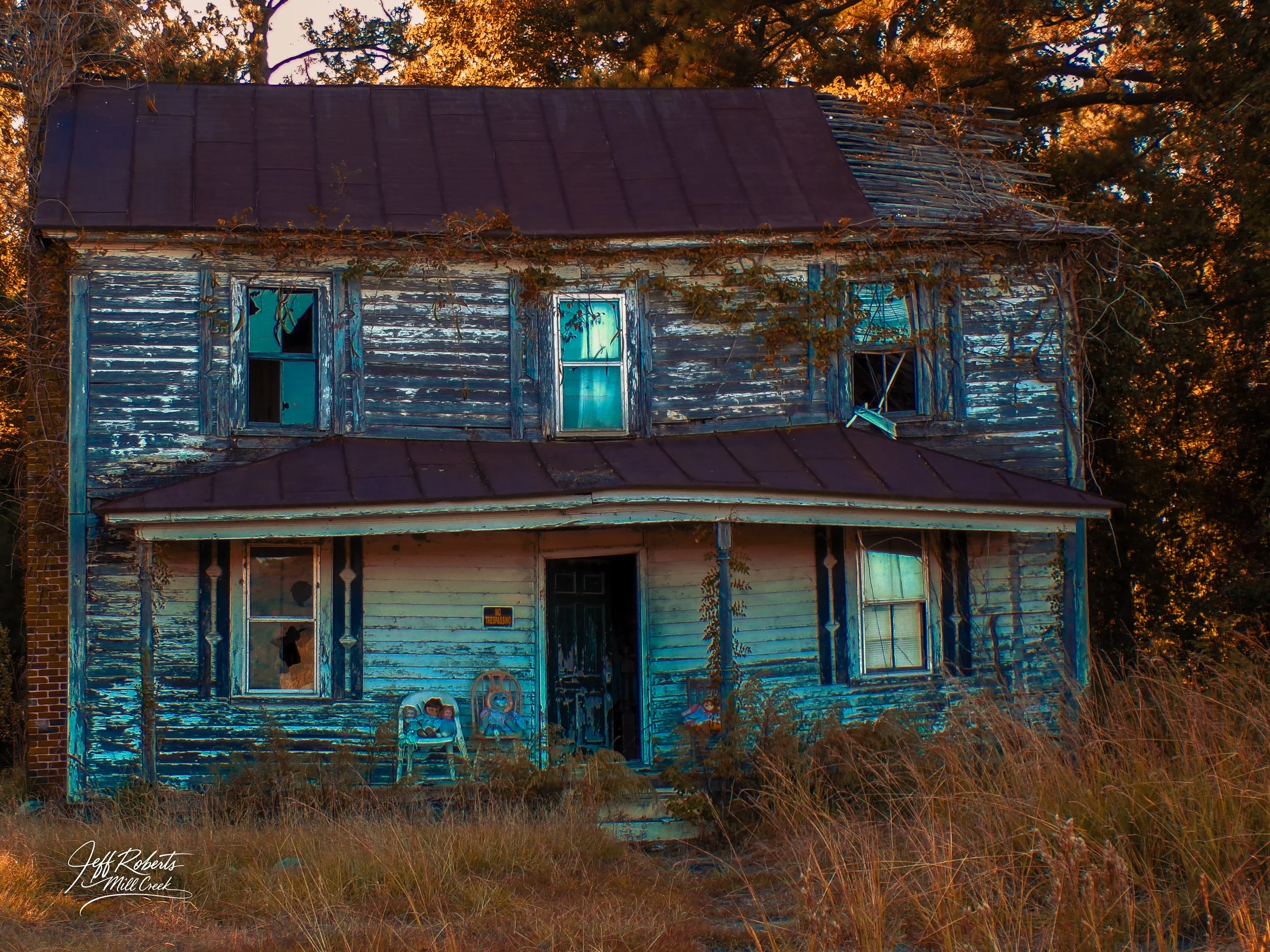 An abandoned two-story house with peeling blue and white paint, broken windows, and a rusty roof, surrounded by tall grass and trees during sunset.