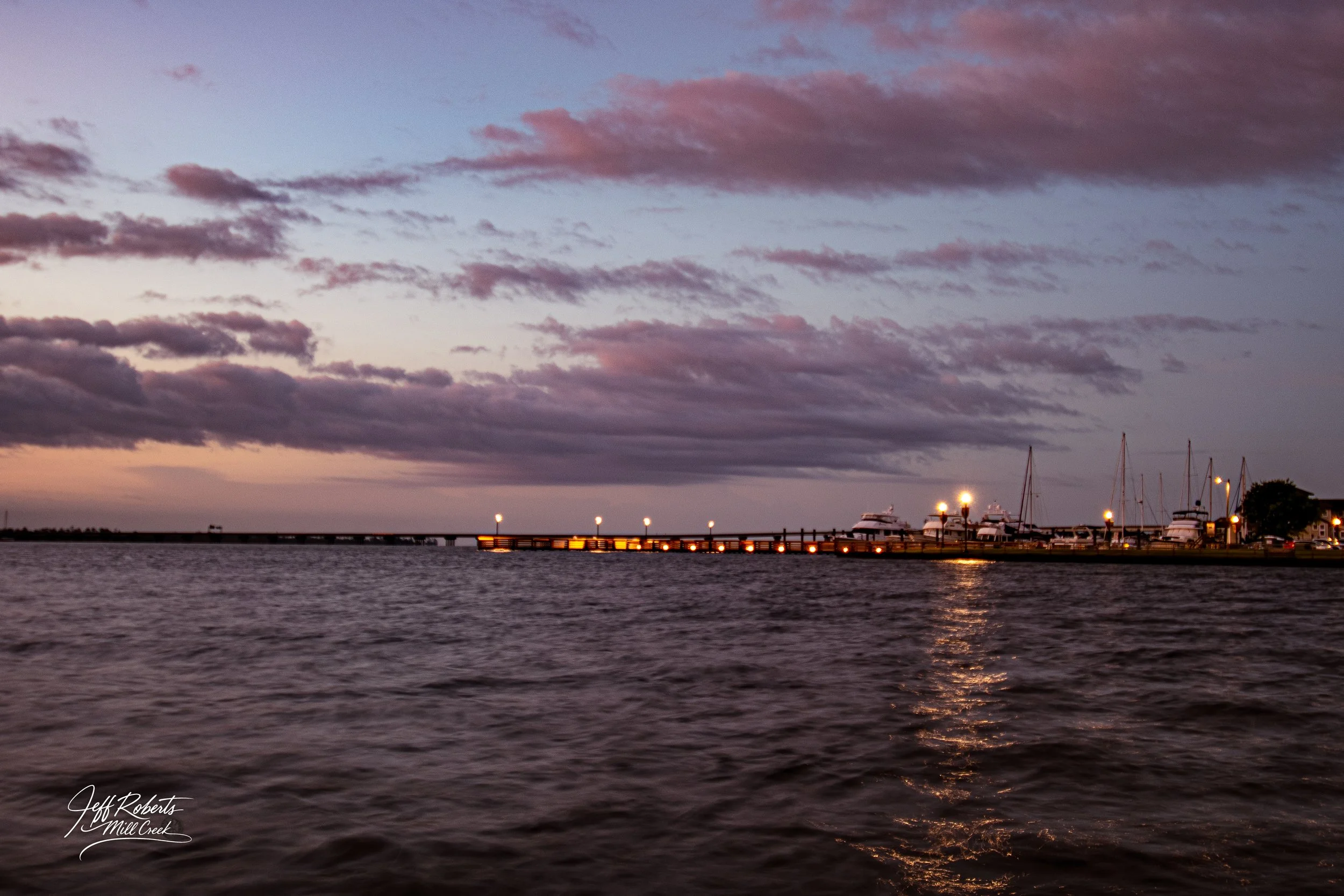 A waterfront dock at dusk with sailboats and yachts, cloudy sky with pink and purple hues, and reflections on the water.