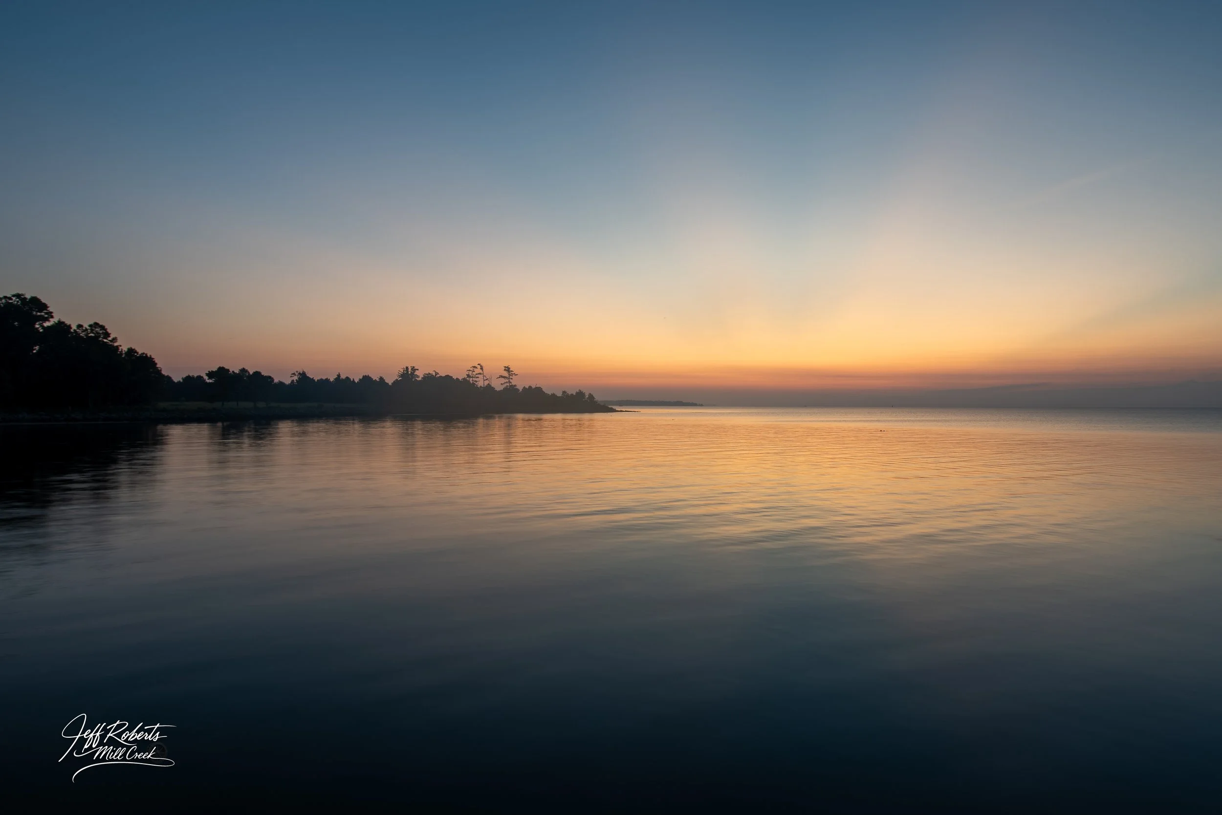 Calm water reflecting a colorful sunset sky with trees on the horizon.