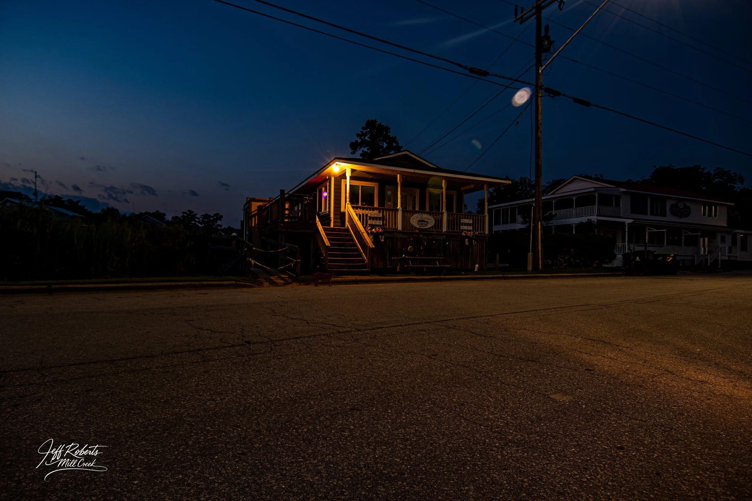 A small, illuminated house on a street at dusk, with a porch, stairs, and power lines overhead.