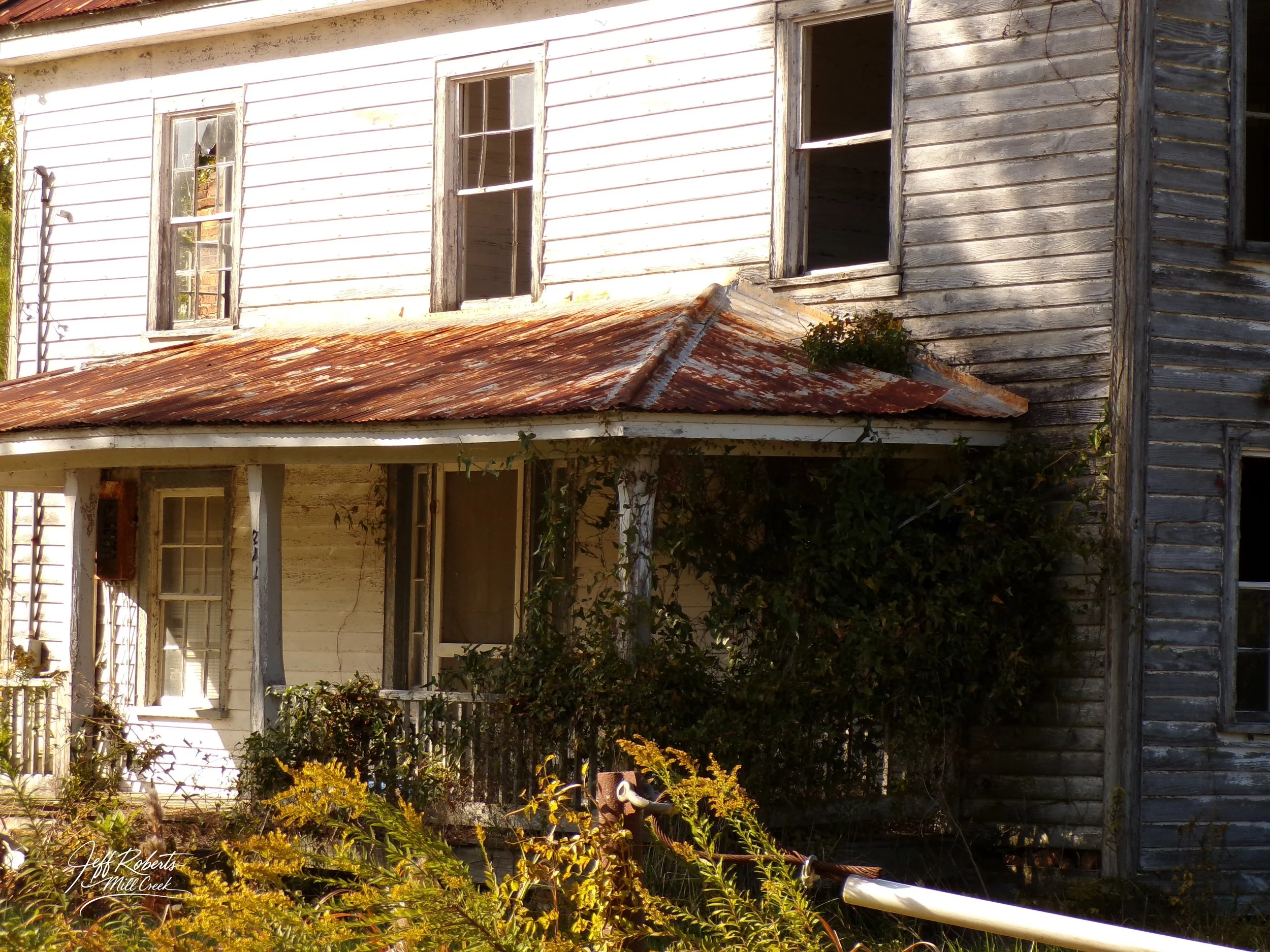 An old, abandoned, two-story wooden house with peeling paint, broken windows, and a rusted metal roof. Overgrown bushes and plants surround the house.