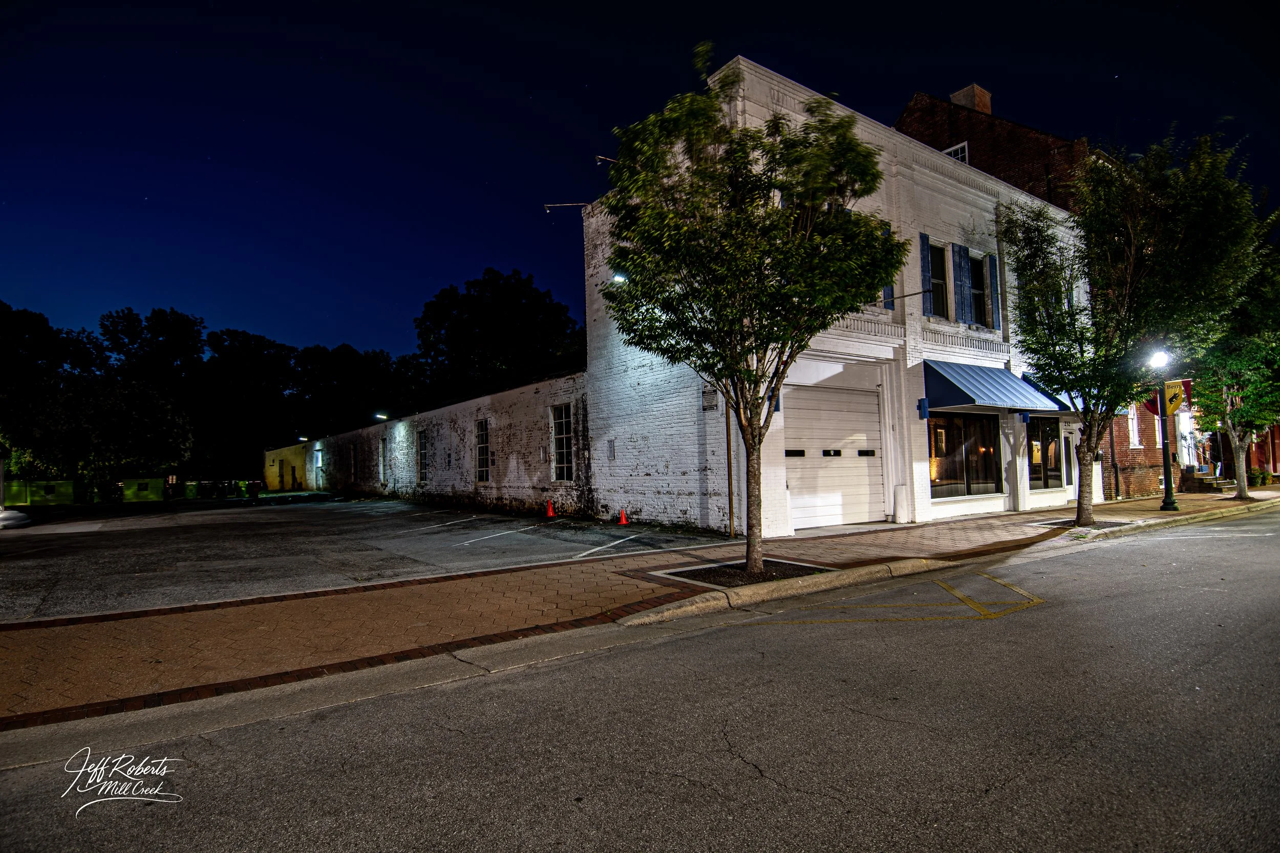 Nighttime view of a white brick building with trees in front, streetlights illuminating the sidewalk, and a dark sky with visible stars.