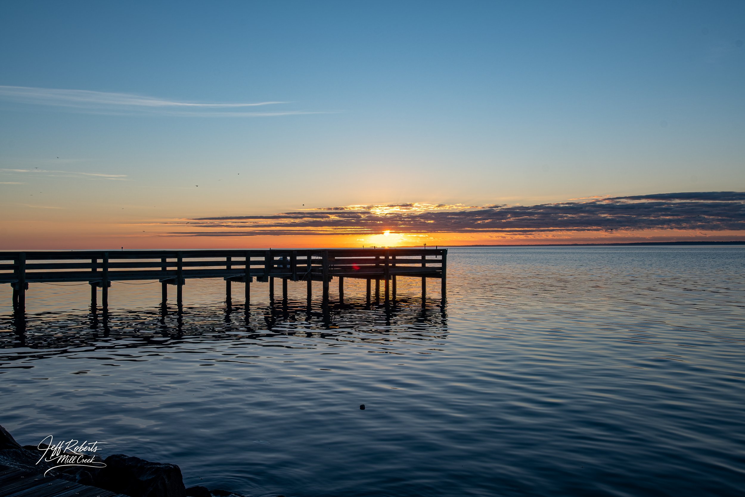 Sunset over a calm lake with a wooden pier extending into the water, and a few birds flying in the sky.