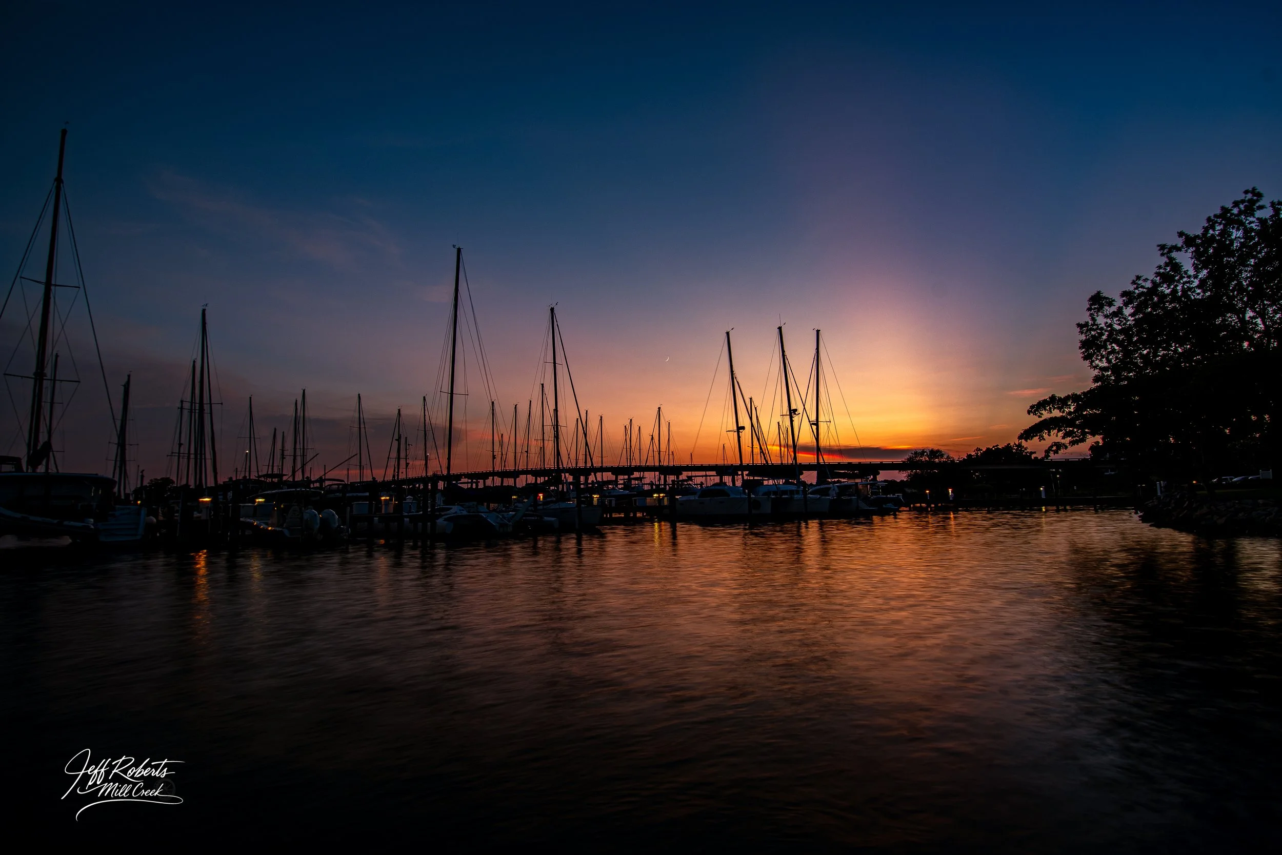 Sailboats docked at a marina during sunset with a colorful sky and calm water reflecting the boats and sky. Signature text at the bottom left corner reads 'Jeff Roberts, Mill Creek.'