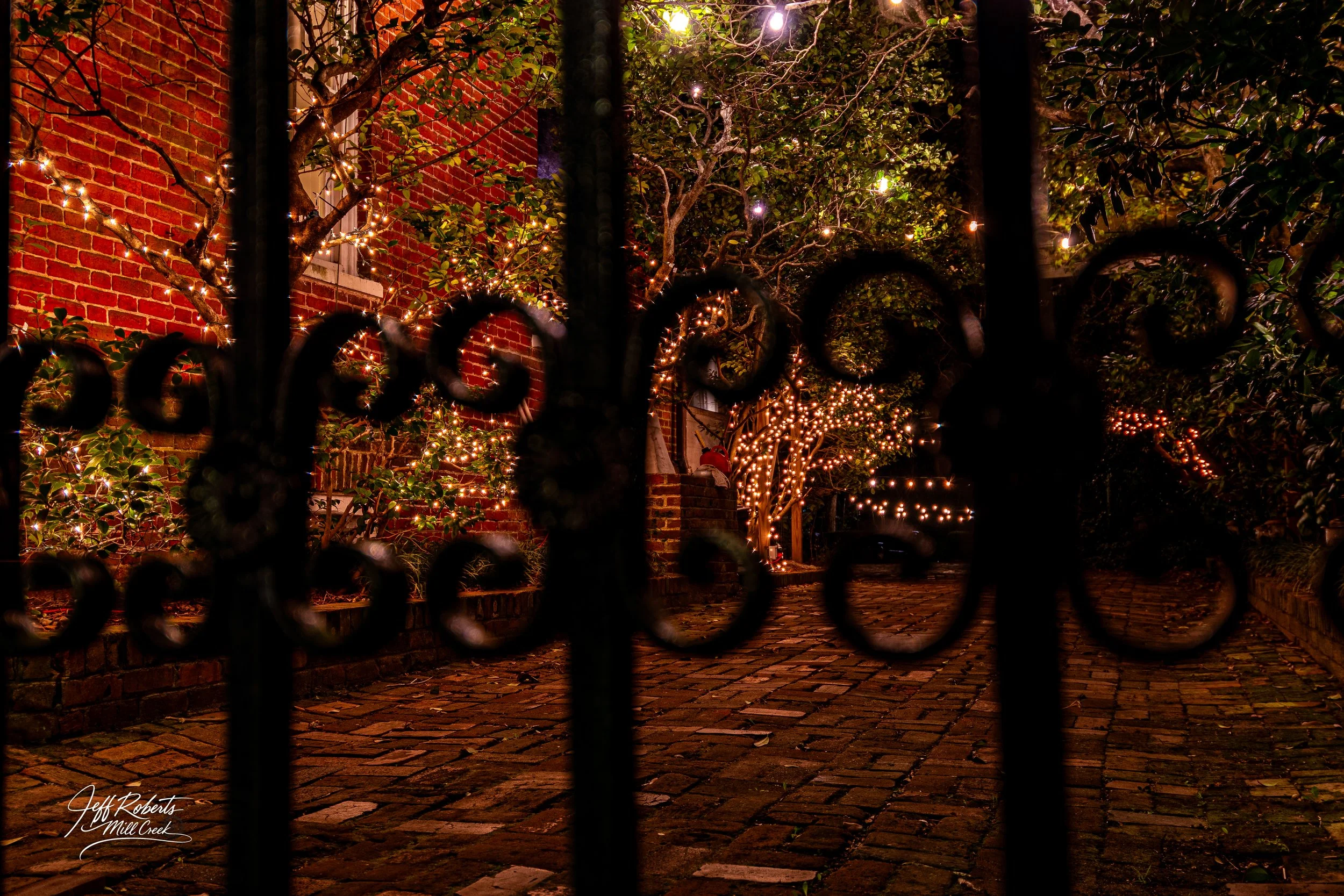 Night view of a brick alleyway decorated with string lights on trees, viewed through a black wrought iron gate.