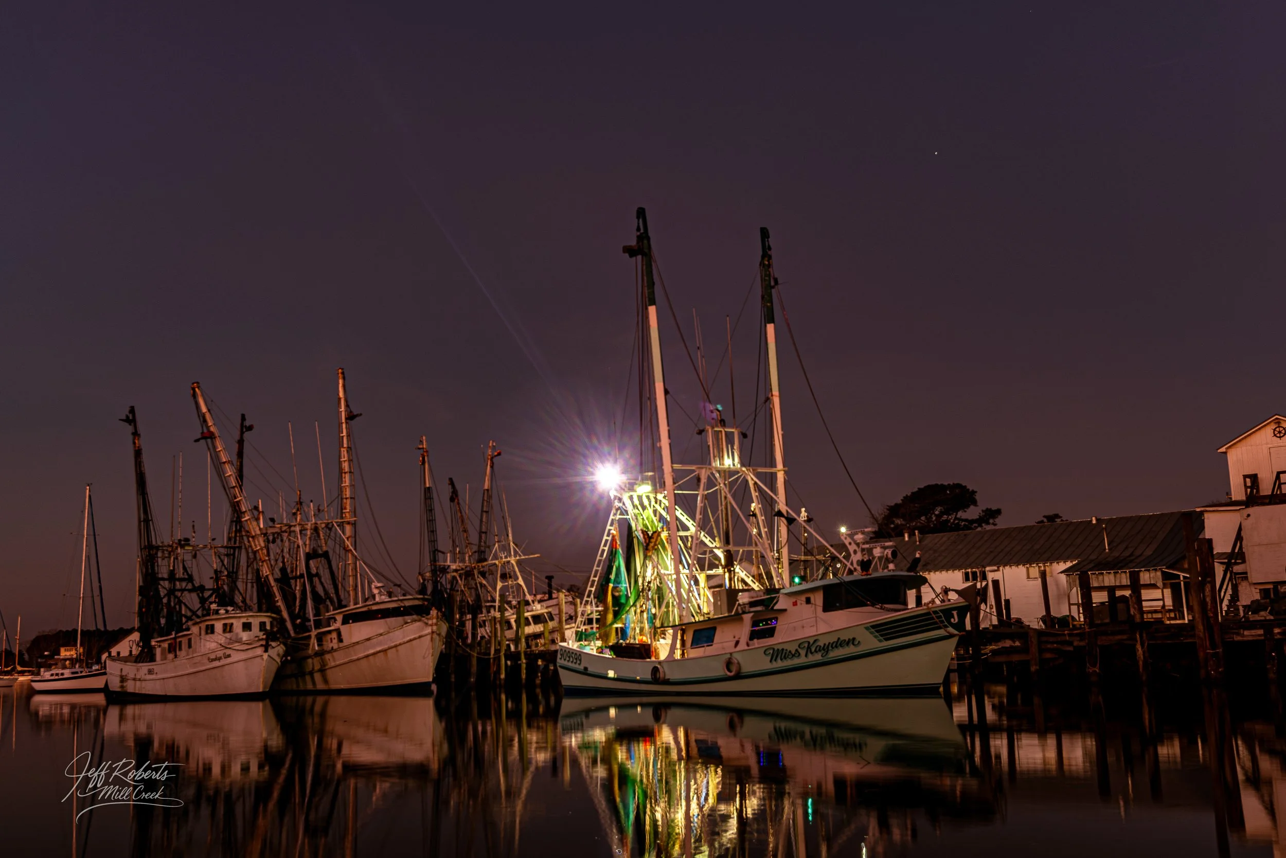Nighttime scene of sailboats docked at a marina, with one boat brightly lit and reflected in calm water, and a clear sky with a visible star.