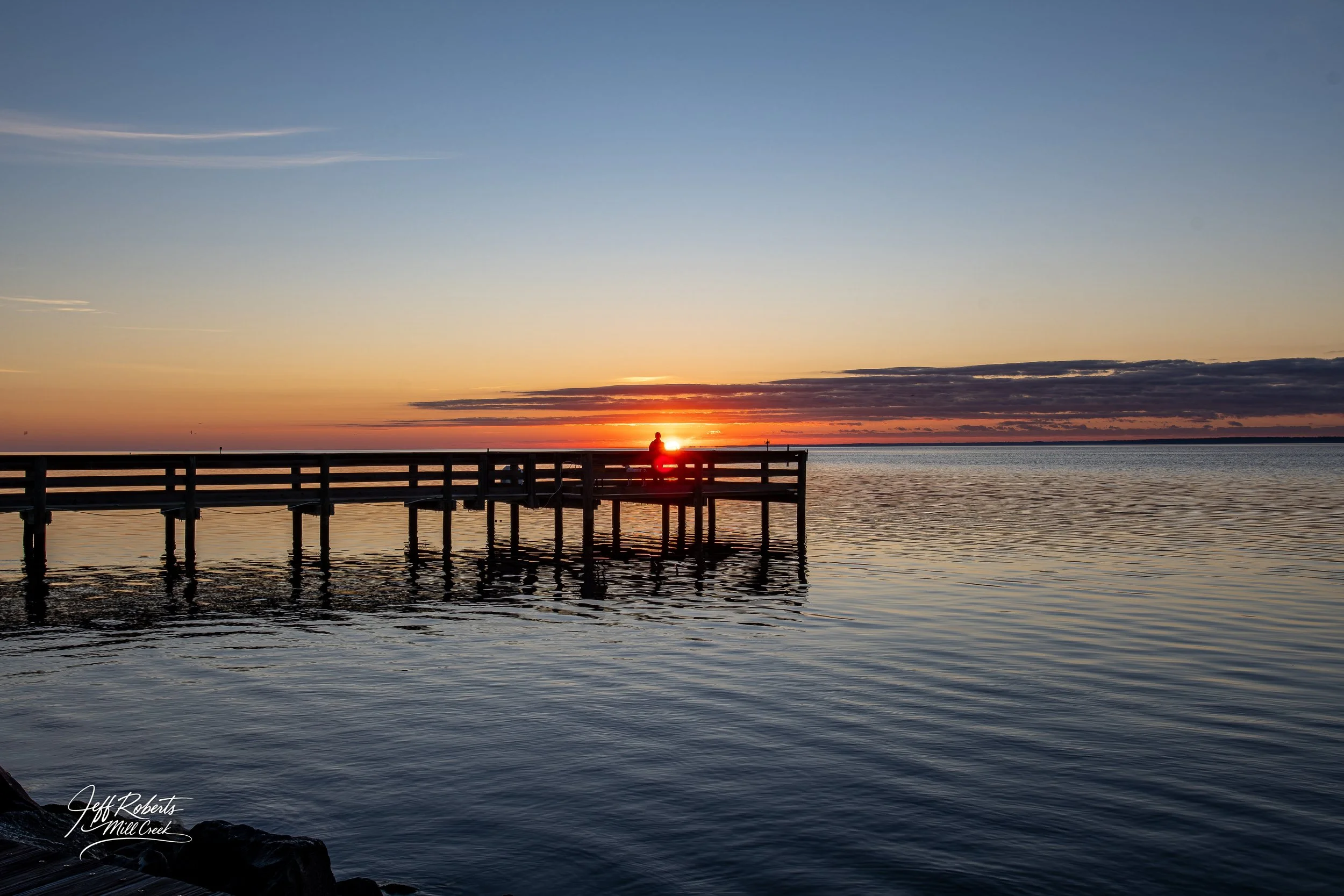 Sunset over a lake with a wooden pier and a person sitting at the end of the pier, silhouetted against the vibrant sky.